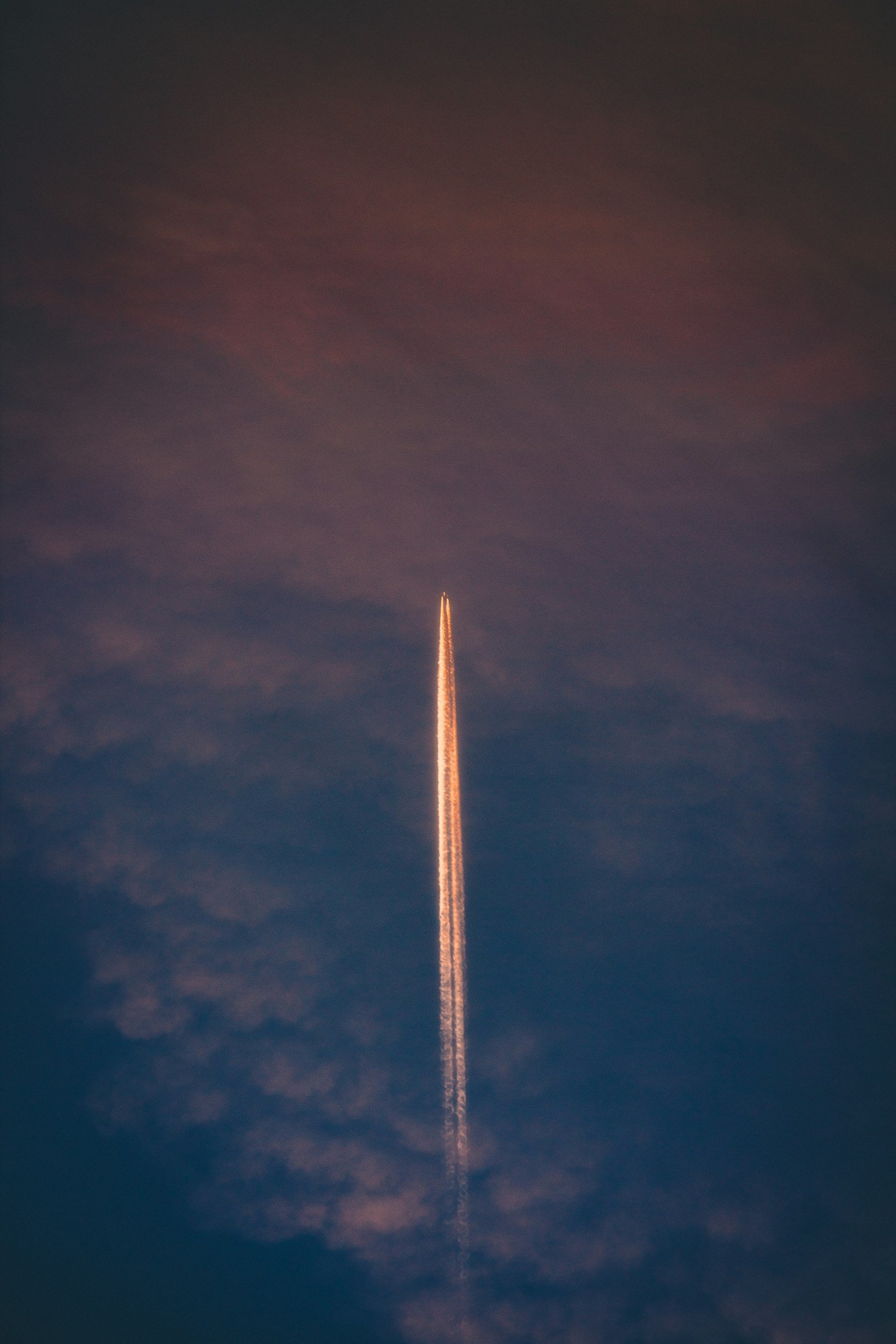 A rocket launches into the sky, leaving a trail of bright light and smoke against a dark evening sky with some clouds.