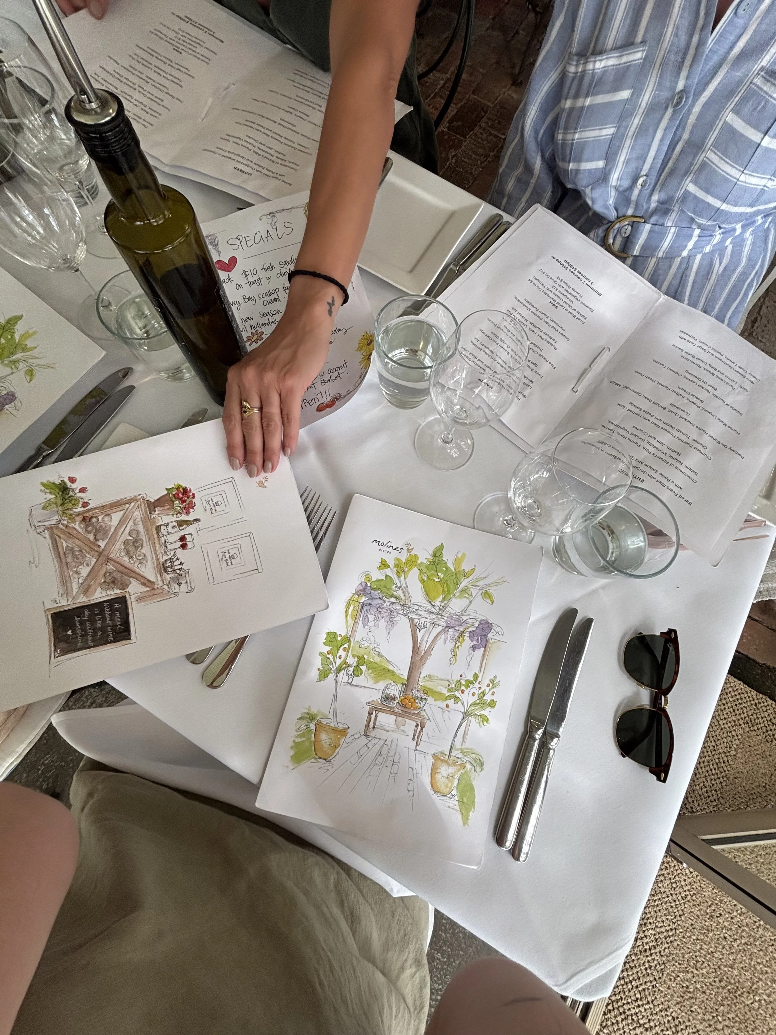 A dining table is set with glasses, silverware, and illustrated menus, with a person reaching for a bottle of olive oil or wine. Hand-drawn place cards or art are also on the table.