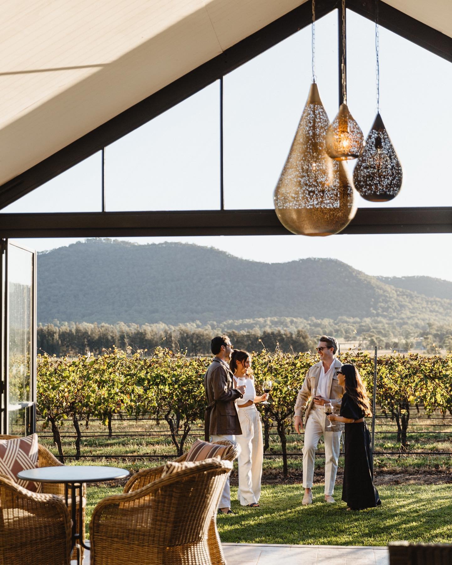 Group of four people standing outside a vineyard in the late afternoon, holding wine glasses and socializing, with rolling hills in the background and modern hanging light fixtures inside a building.