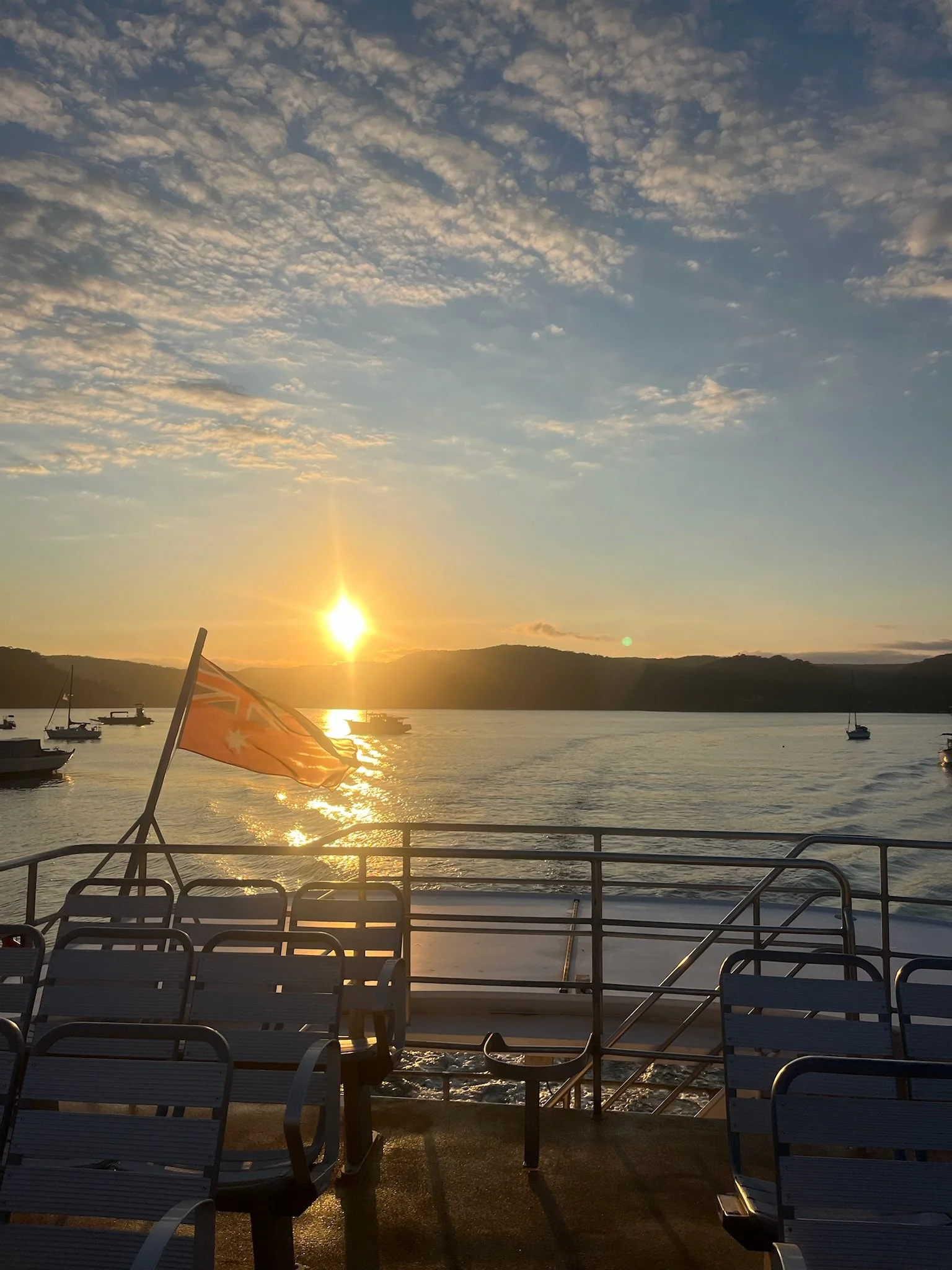 Sunset over a calm body of water with boats, a flag on a boat, and empty chairs on the deck in the foreground.