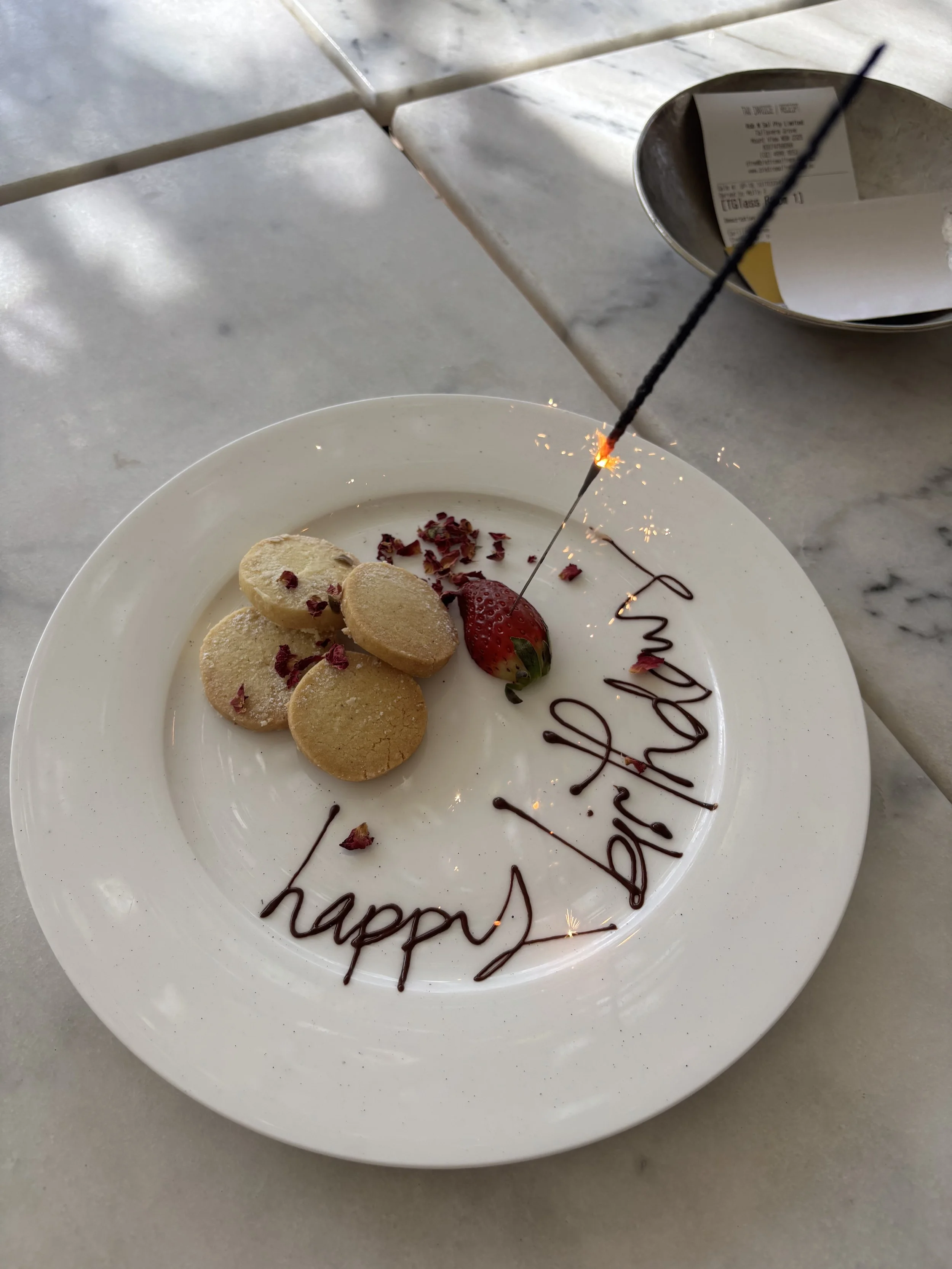 A white plate with cookies, a strawberry, and a birthday message written in chocolate that says 'happy birthday.' A sparkler is lit and burning on the plate, and there is a small metal dish with some receipts or papers in the background on a marble t