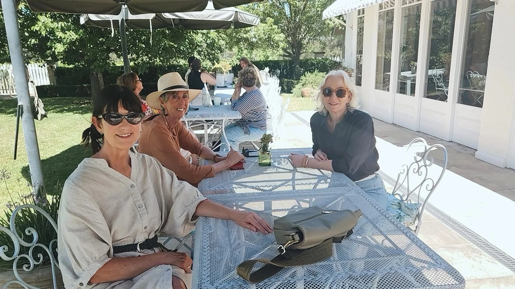People sitting at a long outdoor table under umbrellas on a patio, with a green backyard in the background.