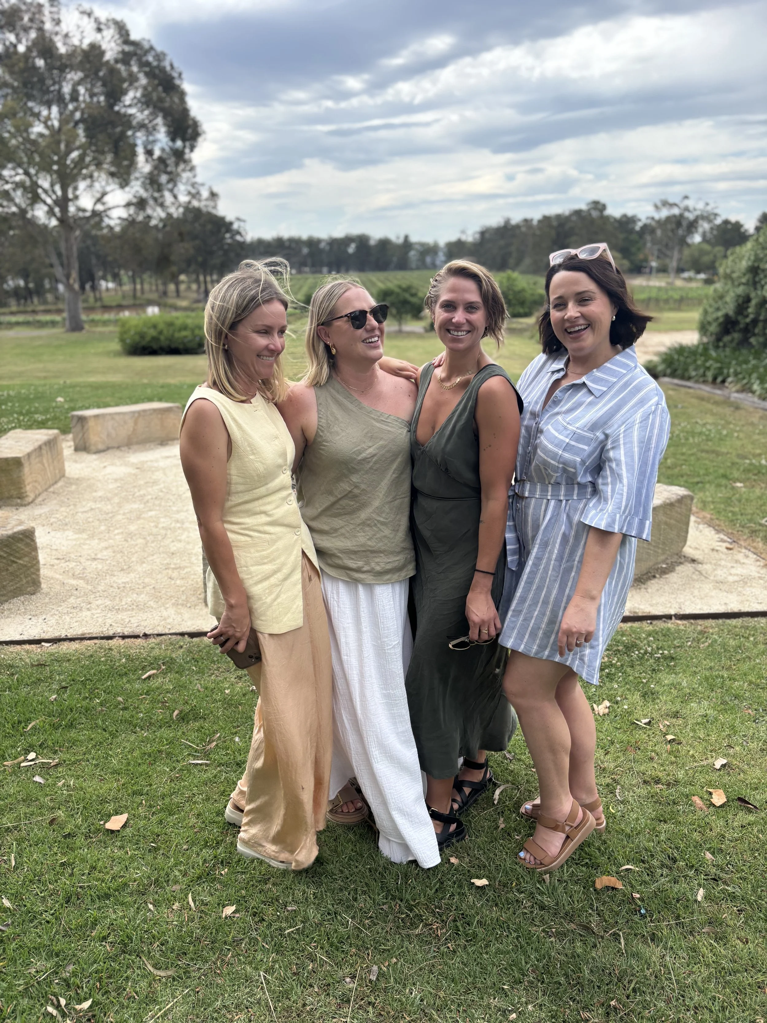 Four women standing outdoors on grass, smiling, with trees and a cloudy sky in the background.