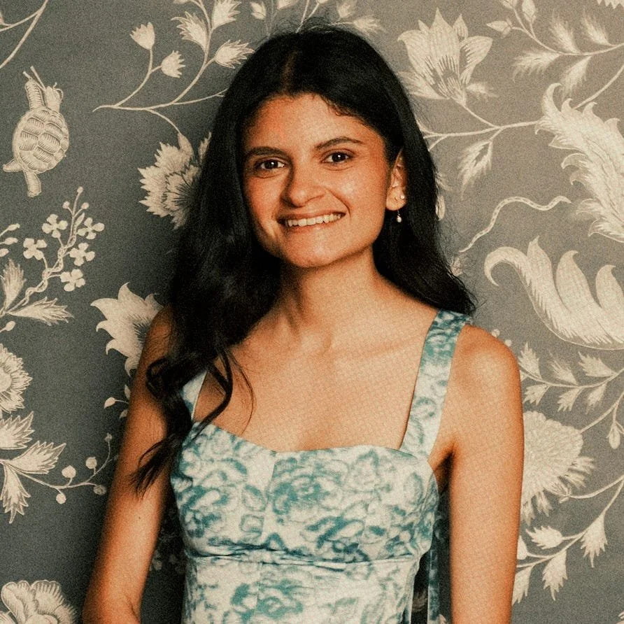 Young woman with dark hair in a floral blue-green dress standing in front of a floral wallpaper background.