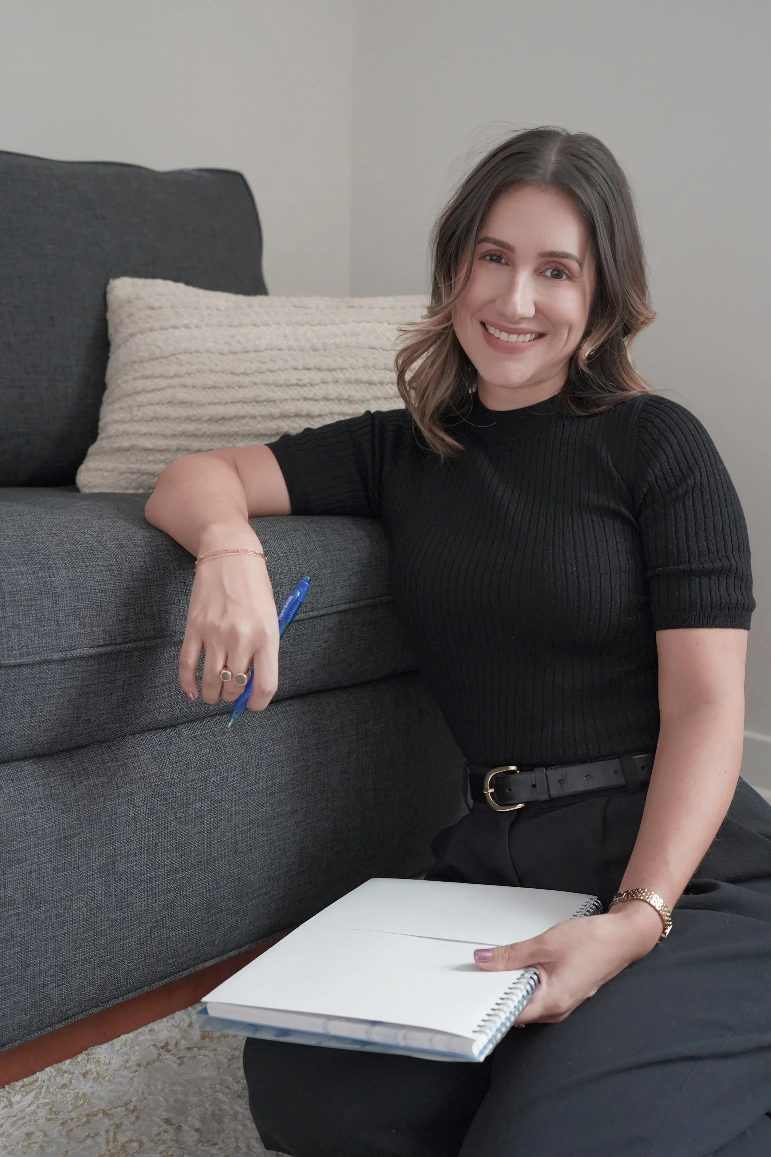 A woman with shoulder-length brown hair, wearing a black ribbed shirt and a gold watch, sitting on the floor next to a gray couch. She is smiling at the camera, holding a white spiral notebook and a blue pen. There is a white knitted cushion on the couch behind her, and a carpet is visible underneath.