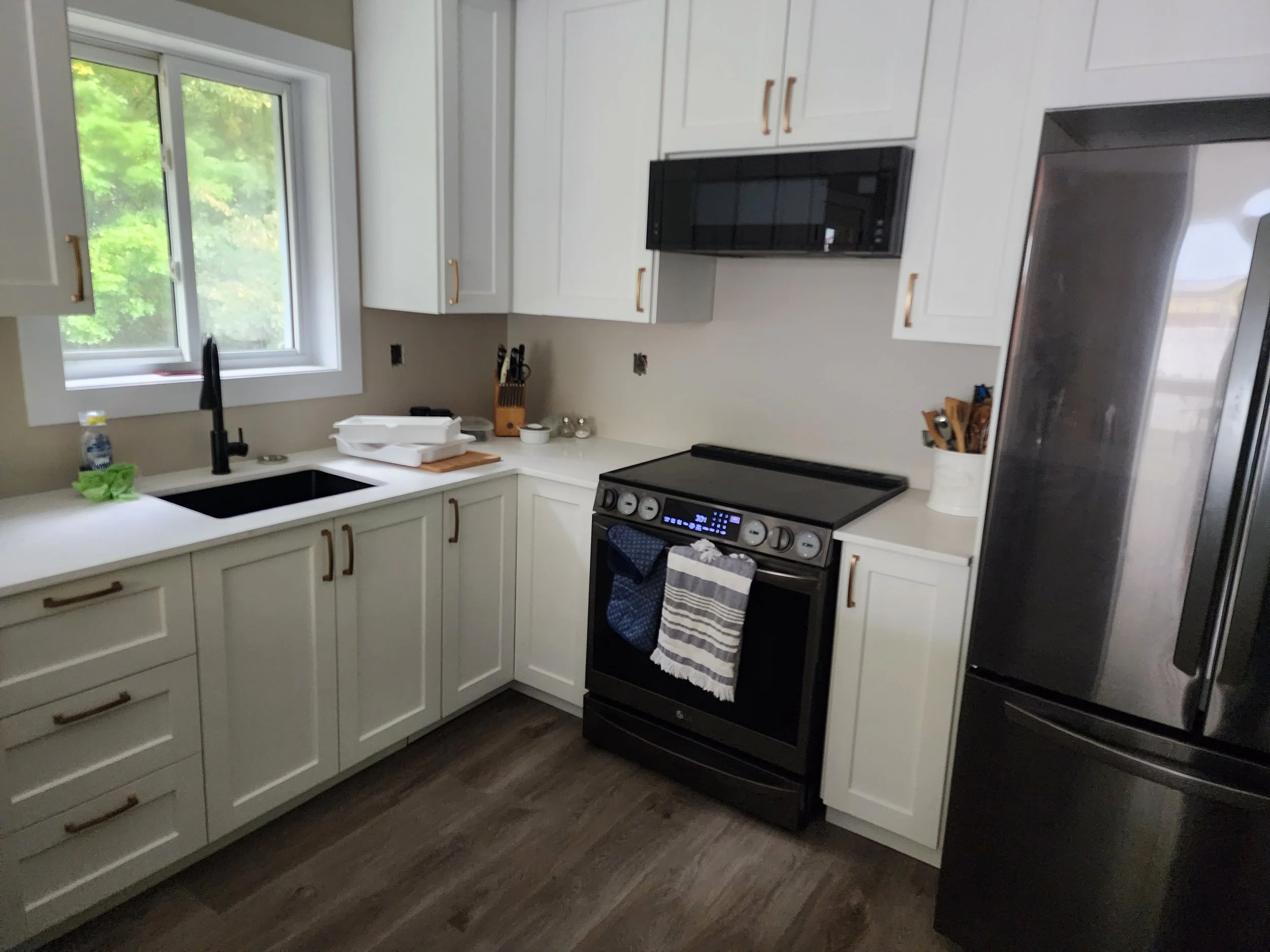 Kitchen with white cabinets, black sink, stove, microwave, and a stainless steel refrigerator. There is a window above the sink with greenery visible outside.