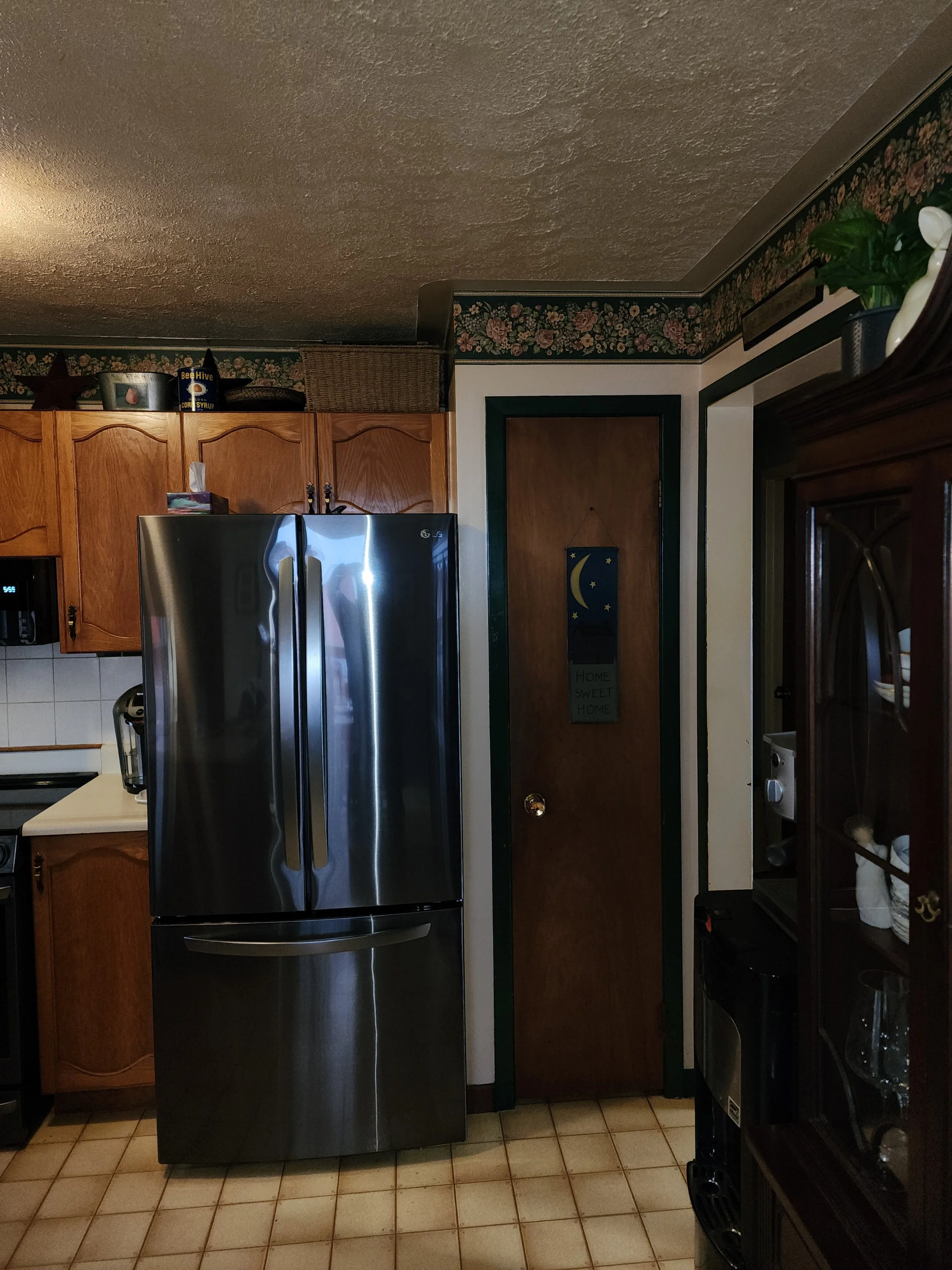 A kitchen with wooden cabinets, a stainless steel refrigerator, a door with a moon and star decoration, and floral wallpaper border.