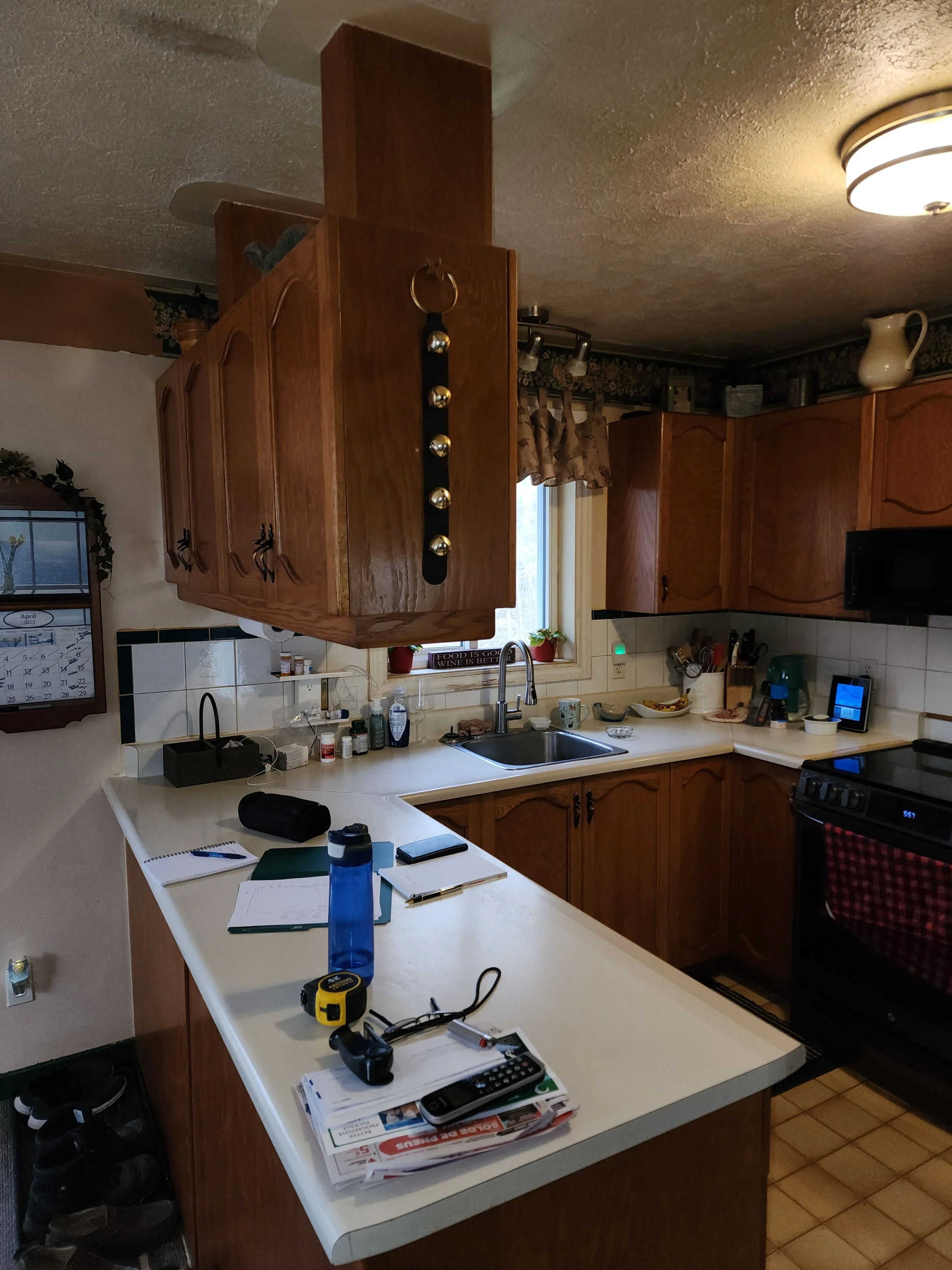 Kitchen with wooden cabinets, a white countertop, a window with a plant, and various items on the counter, including notebooks, a water bottle, and a remote control.