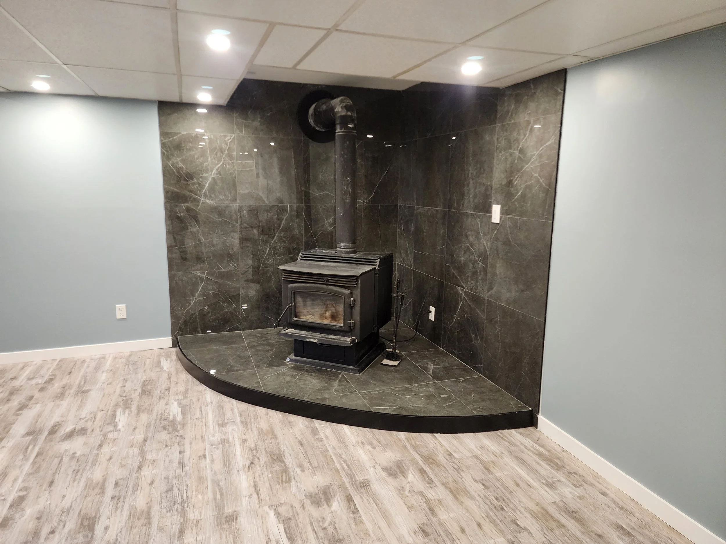 A room with a black wood stove set on a dark marble hearth, surrounded by dark marble walls, with light blue painted walls on the sides, wood-like flooring, and ceiling lights.