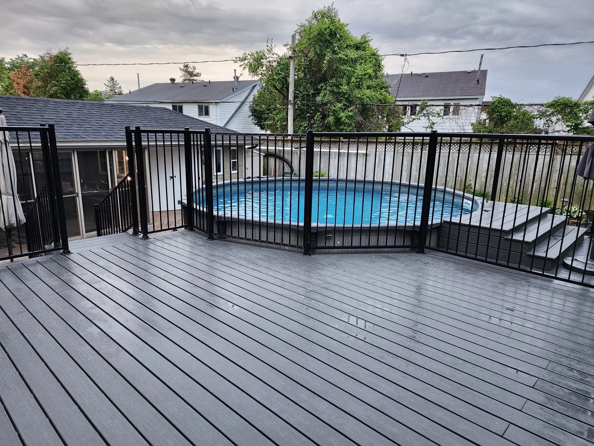 A backyard deck with a round above-ground swimming pool, black metal safety fence, and stairs, surrounded by neighboring houses and trees, under an overcast sky.