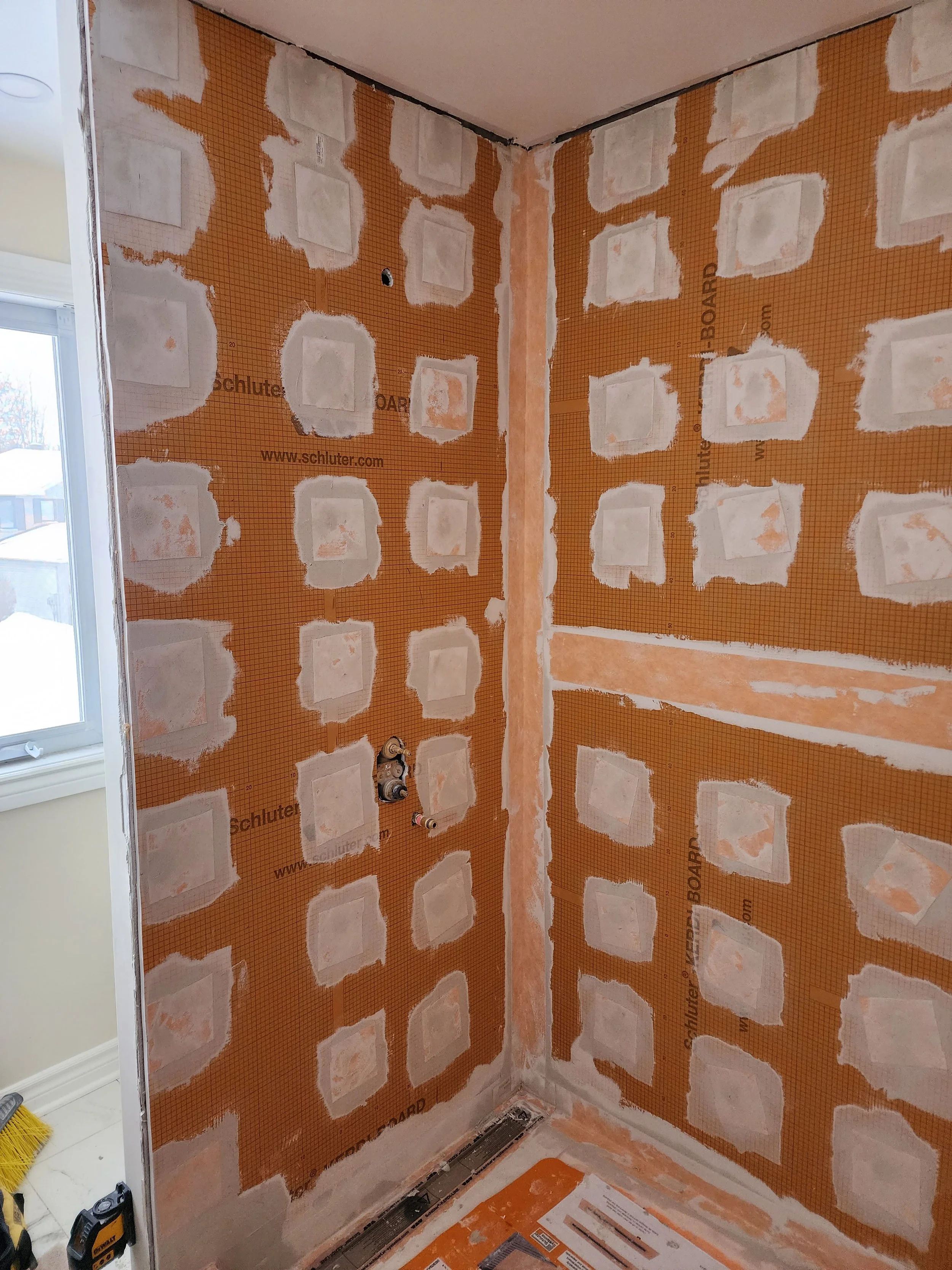 A corner of a room under construction or renovation with drywall panels and joint compound applied, exposed plumbing, and construction materials on the floor.