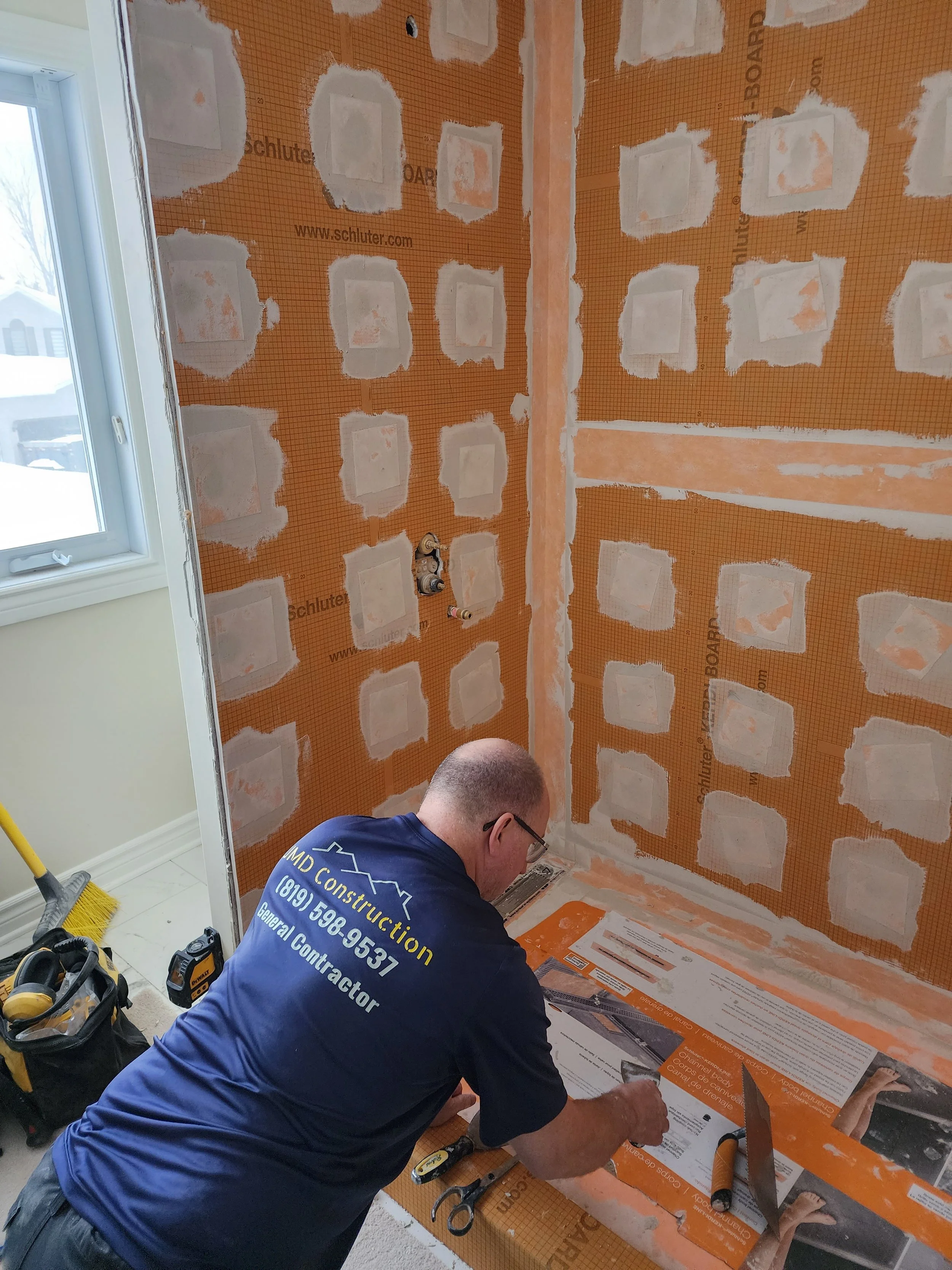 A construction worker kneeling in a bathroom under renovation, working on the floor with tools nearby, surrounded by partially installed wall insulation and drywall. Snow is visible outside the window.