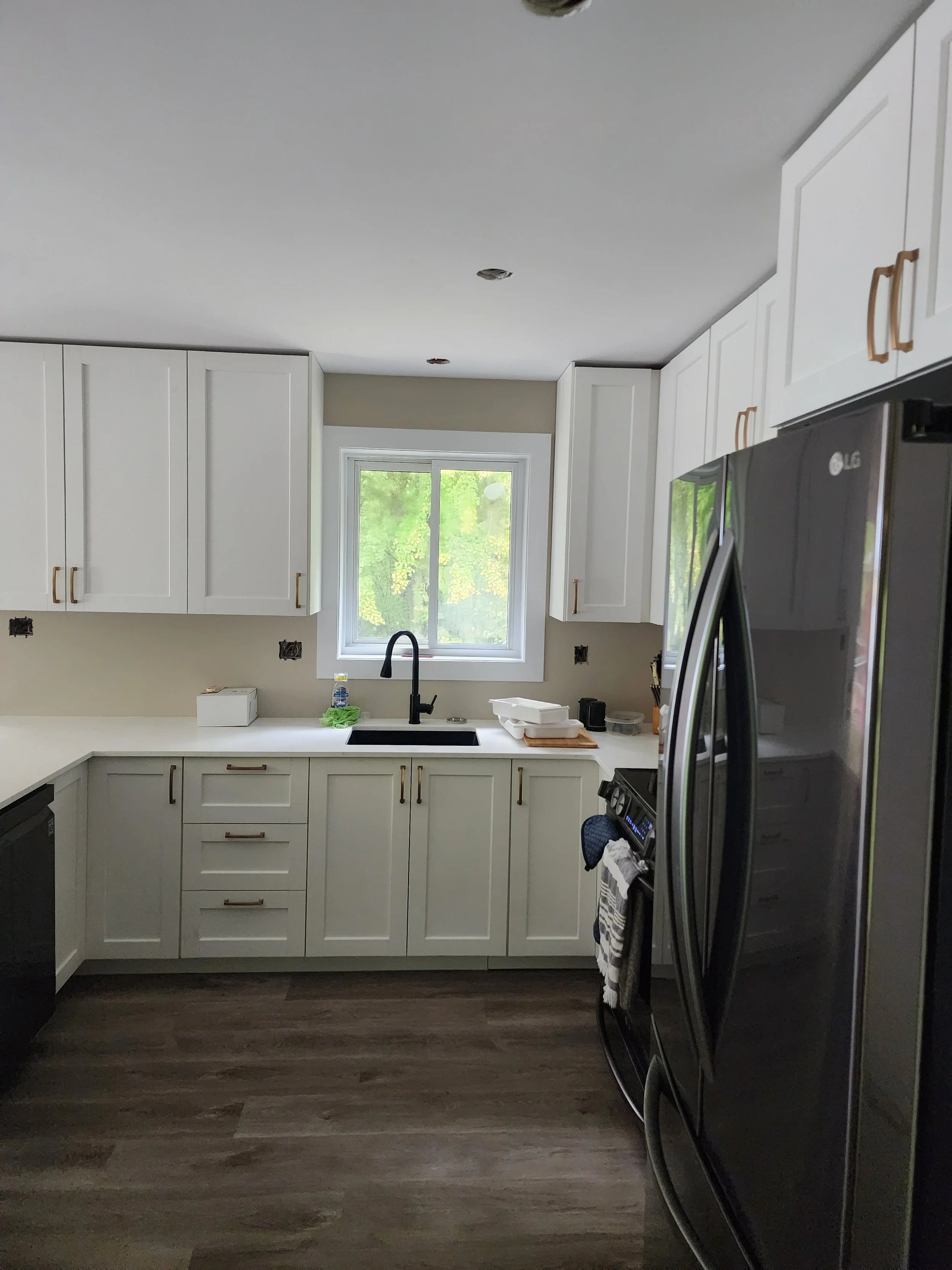 Modern kitchen with white cabinets, black appliances, and a window above the sink looking out to green trees.
