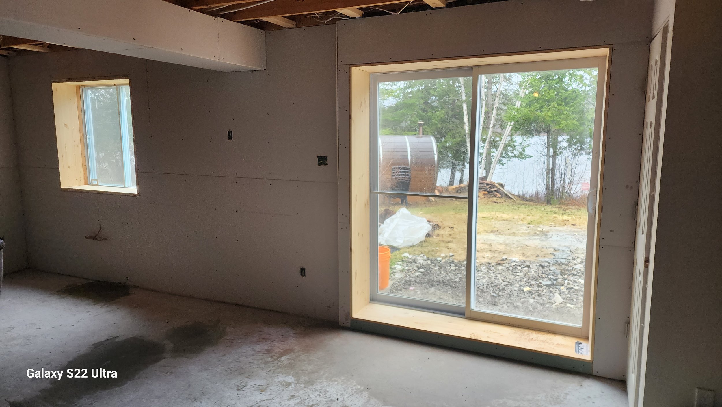 Interior of a house under construction with unfinished drywall, a sliding glass door leading outside to a yard with trees and a lake, and a small window.