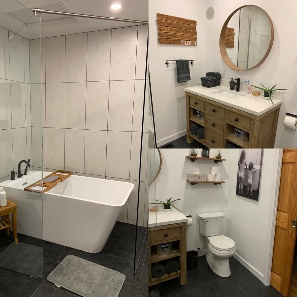 A modern bathroom with a large bathtub, a wooden side table with toiletries, a glass shower enclosure, a wooden vanity with a white countertop, a round mirror, a towel rack, shelves with decorative items, a toilet, and black tiled flooring.