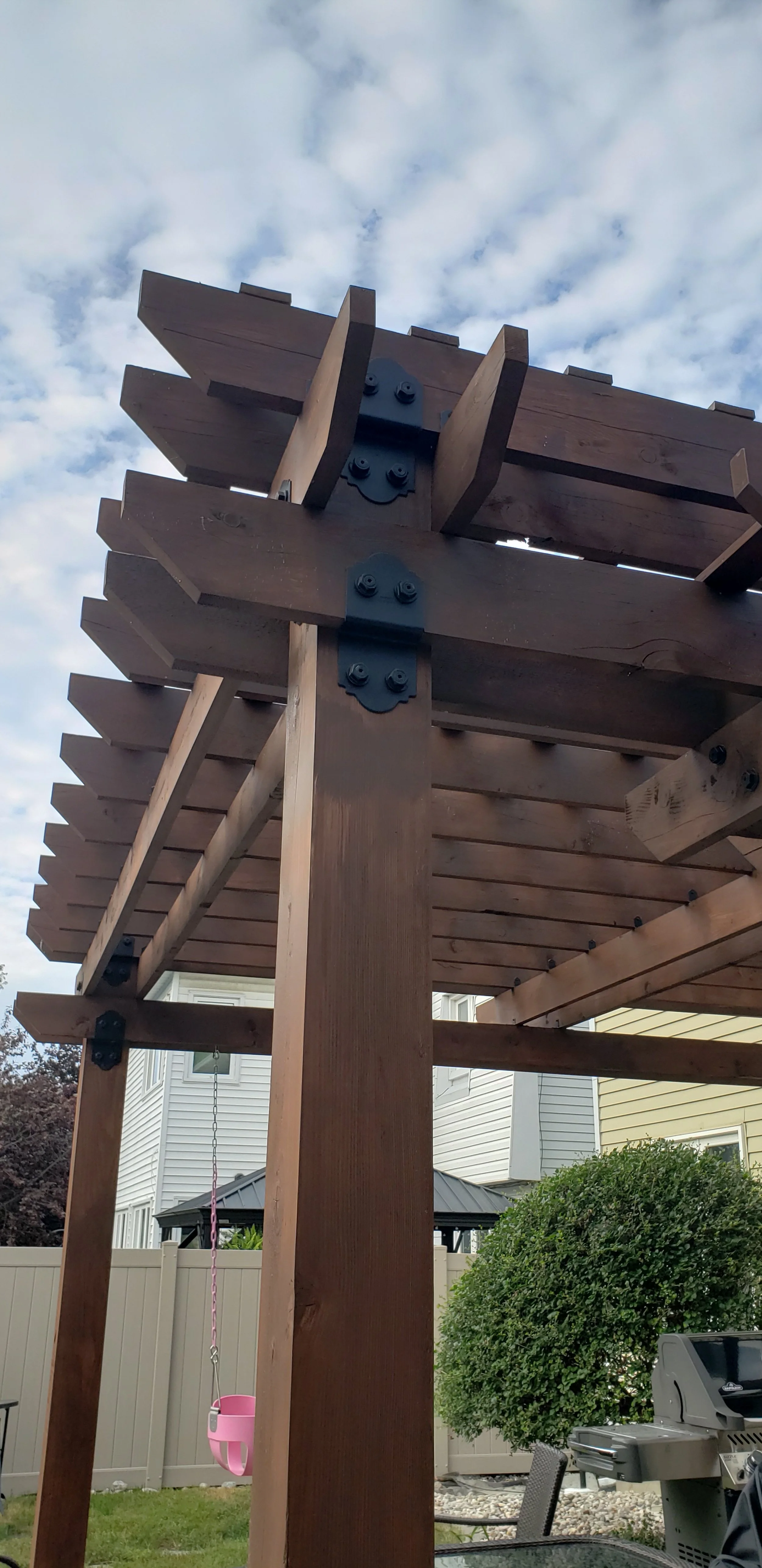 A wooden deck structure under construction in a backyard, with a nearby grill, patio furniture, and a house with siding and a fenced yard, with cloudy sky above.