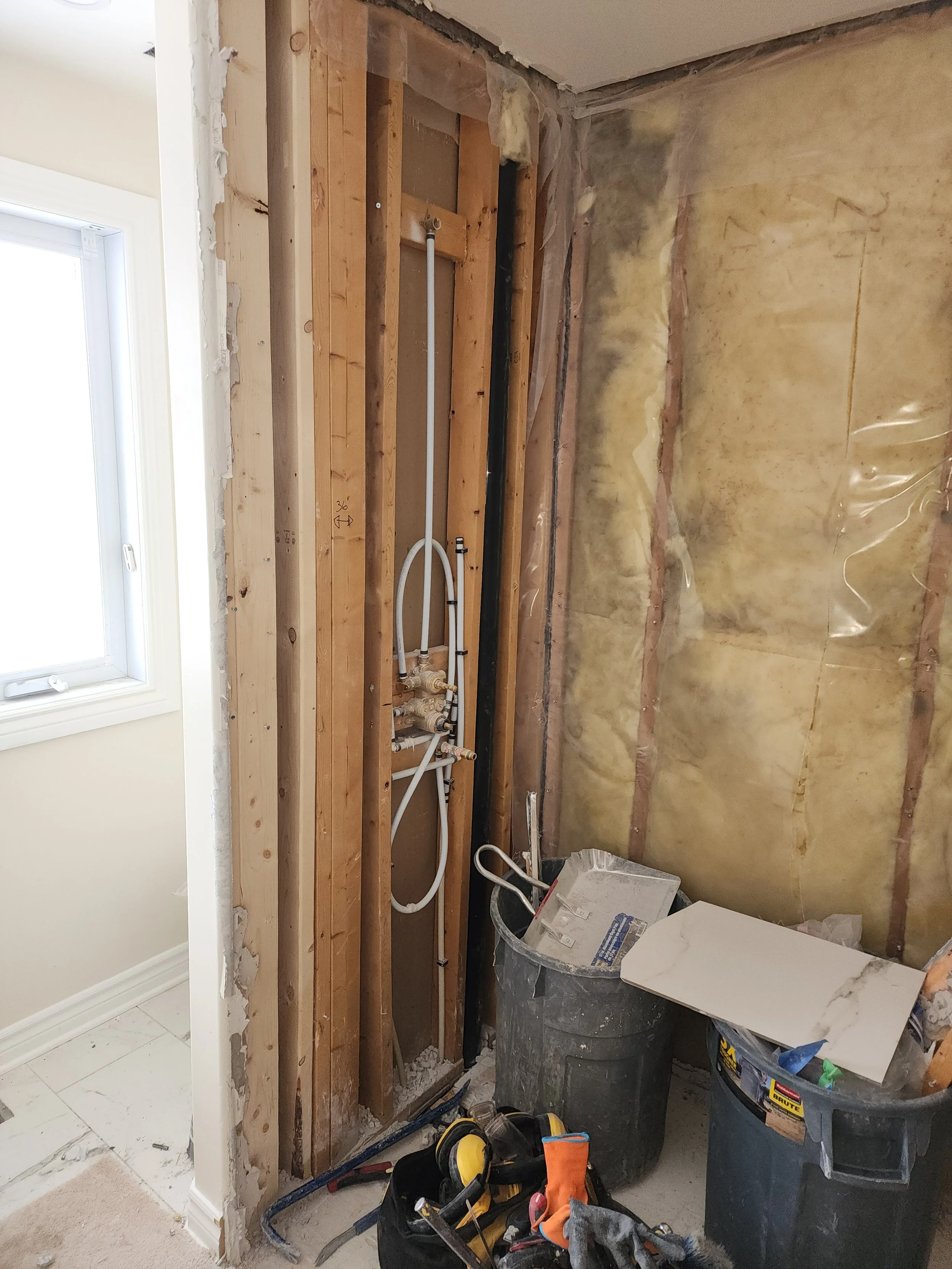 Room under renovation with exposed wall studs, pipes, and insulation, construction tools on the floor.