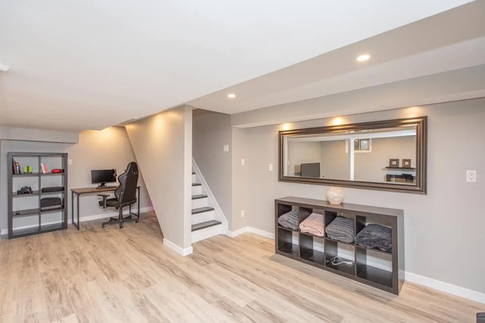 Basement office space with a desk, chair, bookshelf, and a stairway leading upstairs, with light wood flooring and gray walls.