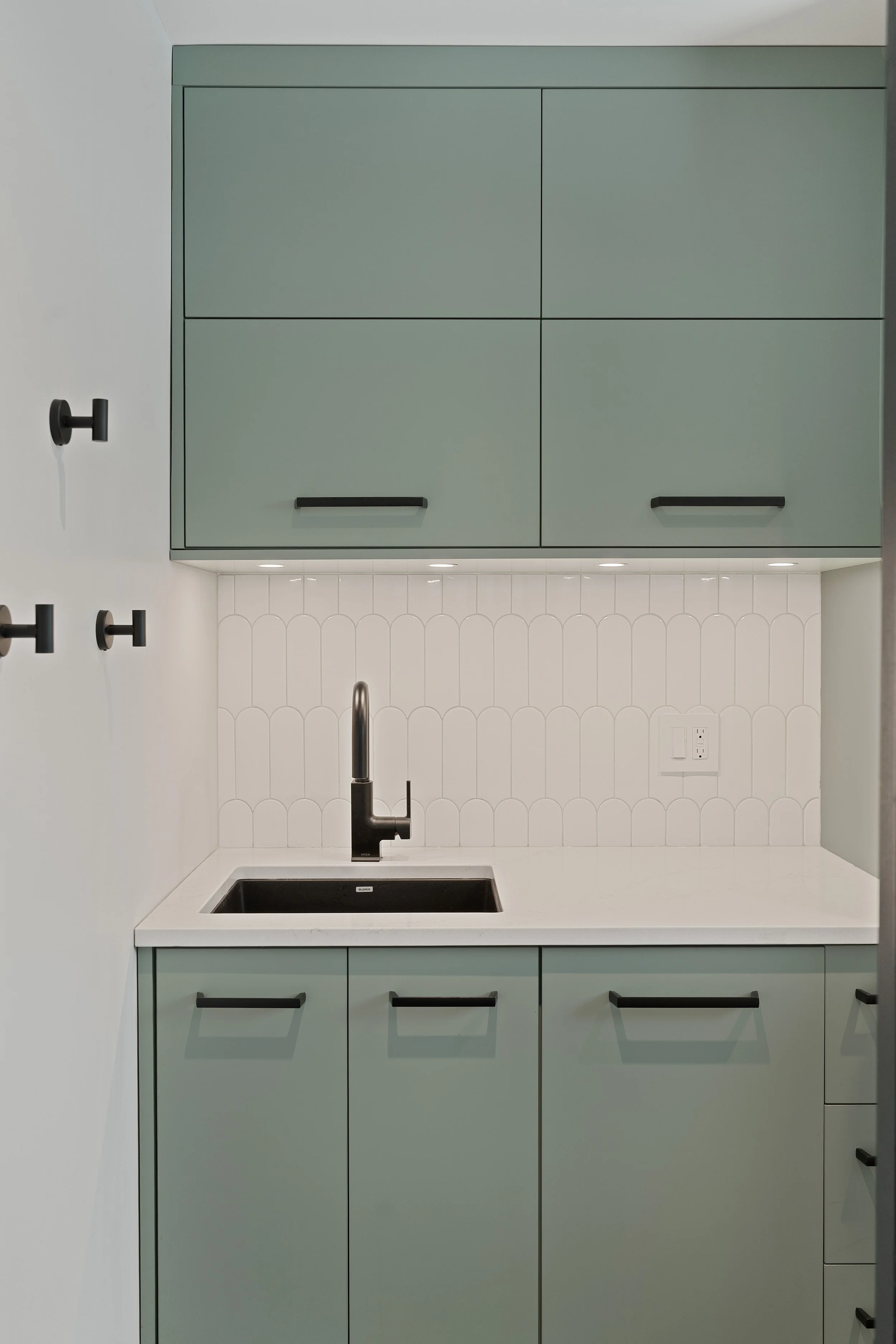 Modern kitchen with mint green cabinets, black handles, white countertops, black sink, and a black faucet. White wall tiles with a scalloped pattern and recessed lighting underneath upper cabinets.