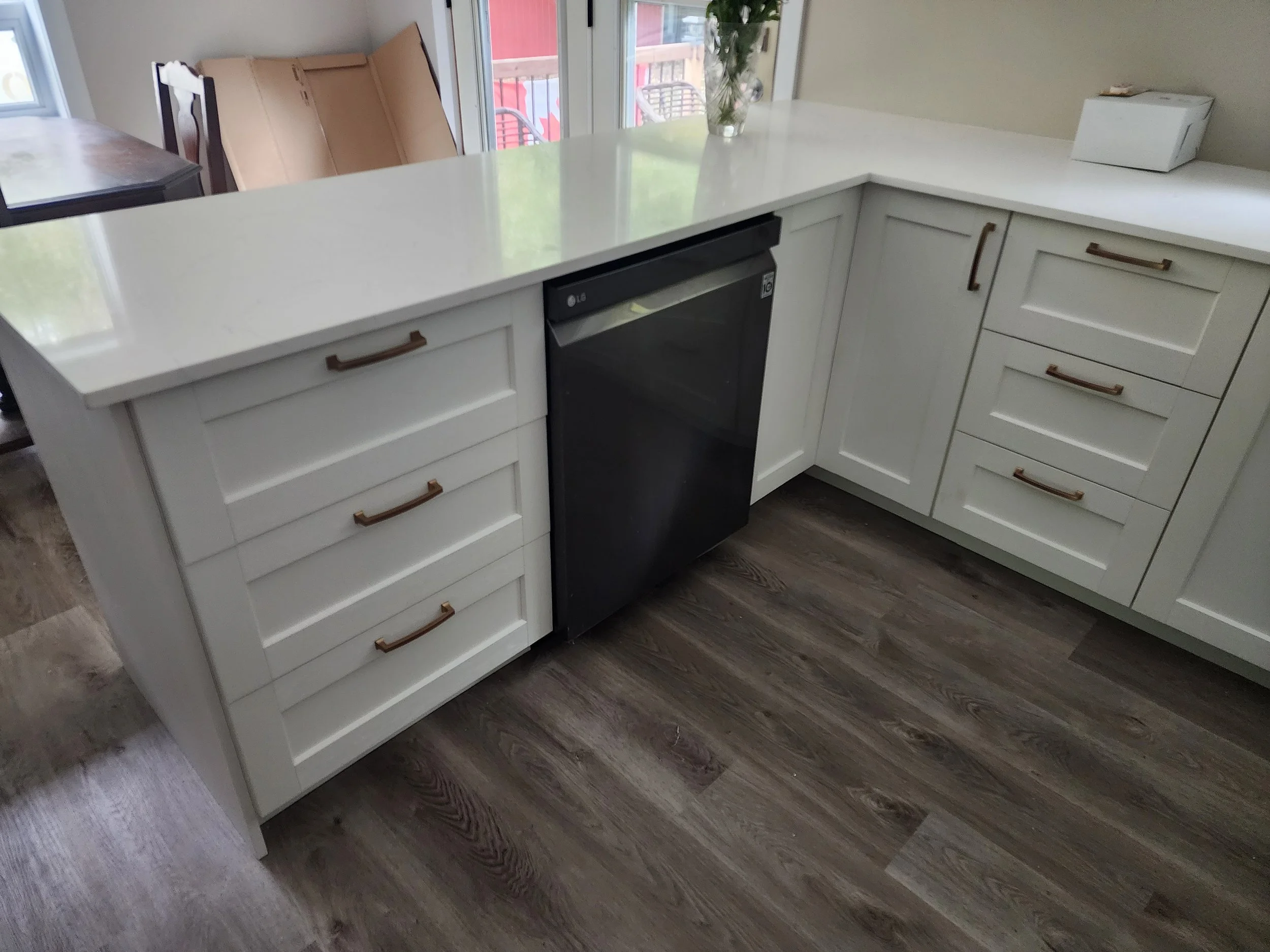 A white kitchen countertop with cabinetry, a black dishwasher, a glass vase with green leaves, and a box on the counter.