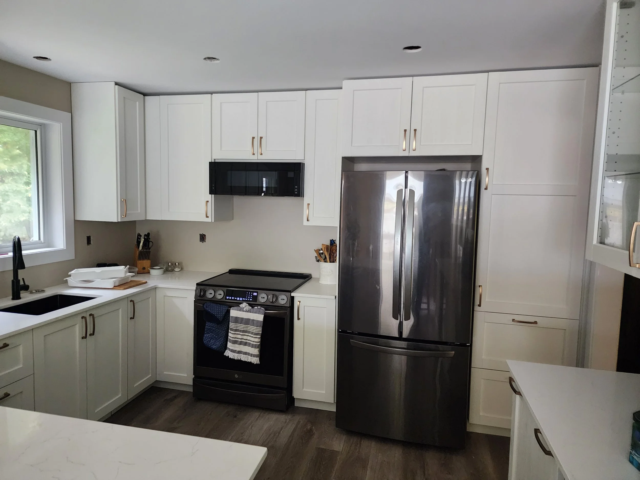 Modern kitchen with white cabinets, black stove, stainless steel refrigerator, black microwave, and black faucet, with a window and wood floor.