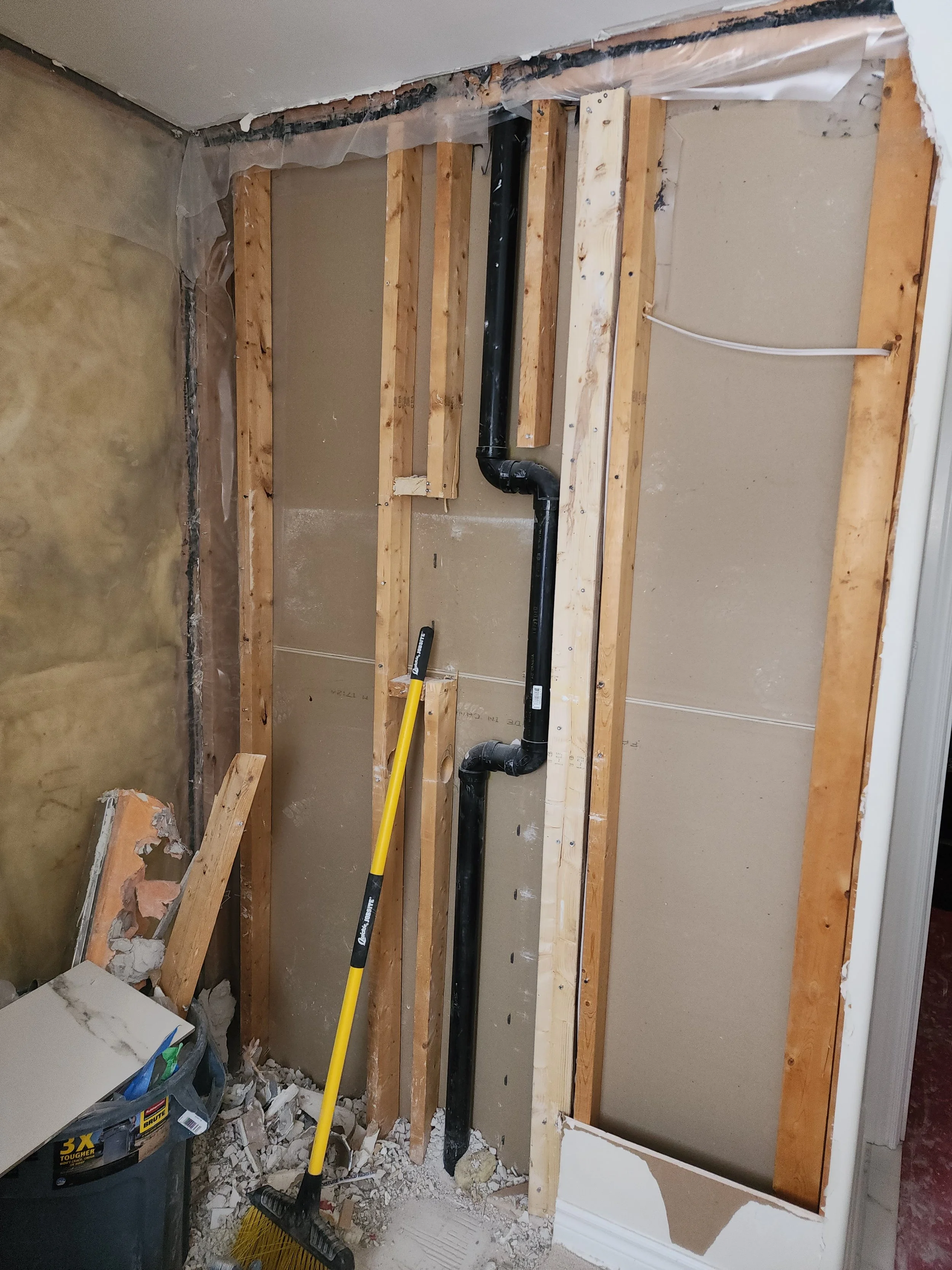 Wall under construction with exposed wooden studs, black plumbing pipes, and a yellow broom leaning against the wall.