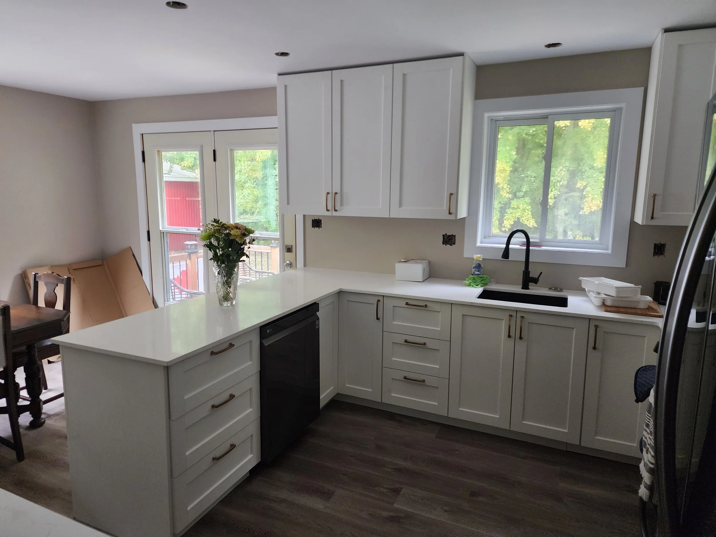 Kitchen with white cabinets, black sink, a window showing green trees outside, and a sliding glass door with a wooden deck outside. A vase with flowers on the counter.