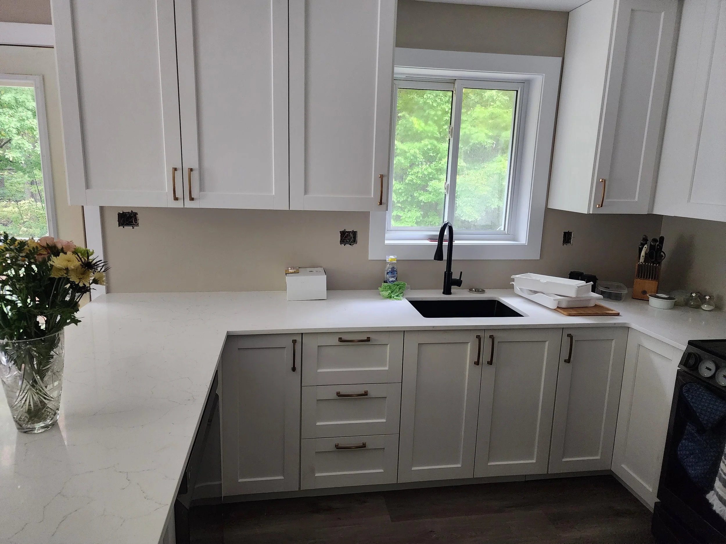 Kitchen with white cabinets, black sink, countertop with flowers and kitchen items, window showing greenery outside.