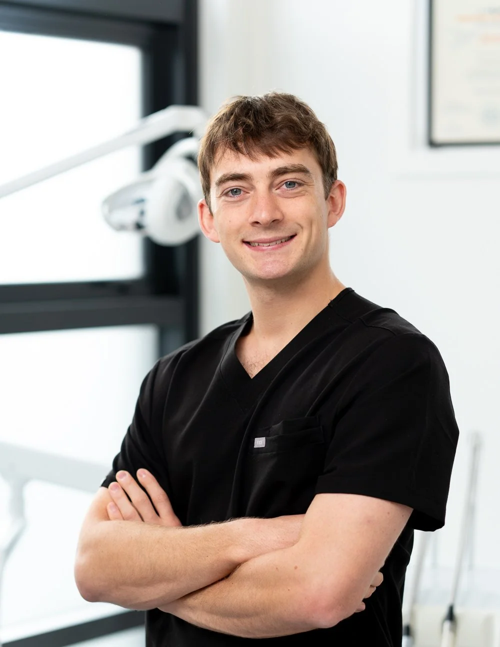 Dr Albert Humphries, a smiling professional dentist in black scrubs standing with arms crossed in a medical office.