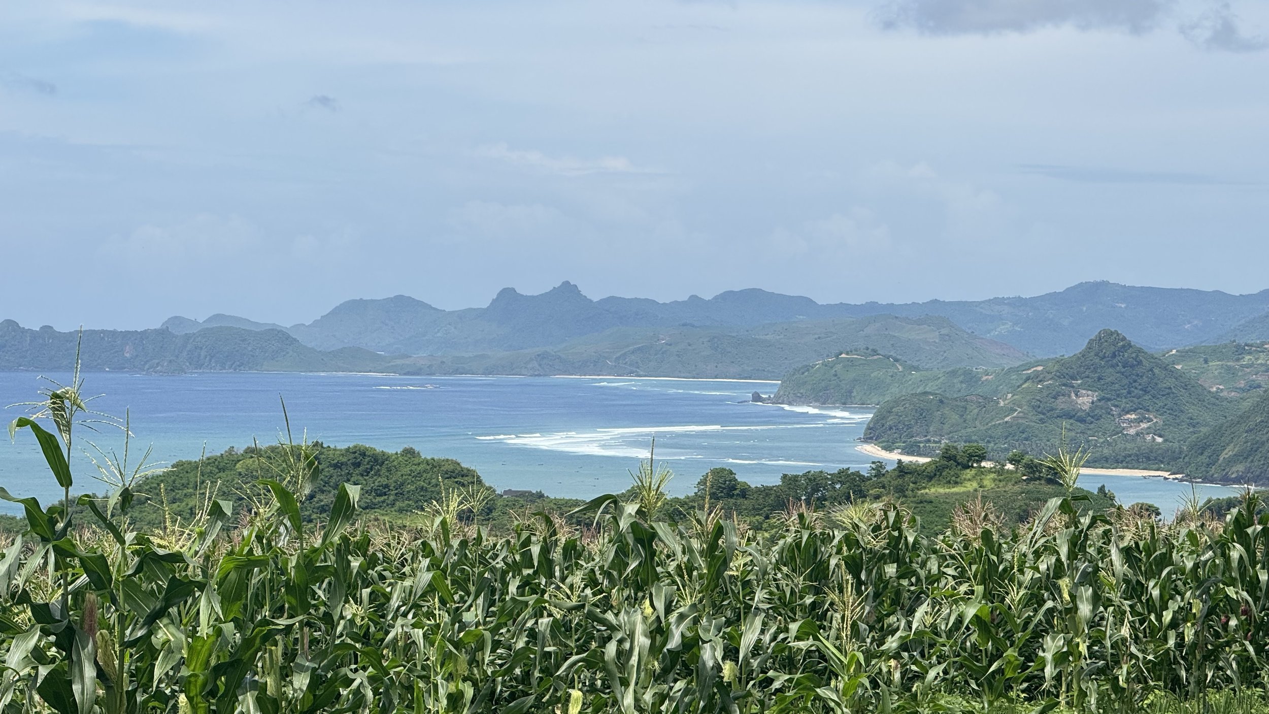 A coastal landscape with green farmland in the foreground, blue ocean waves, and distant mountain ranges under a partly cloudy sky.