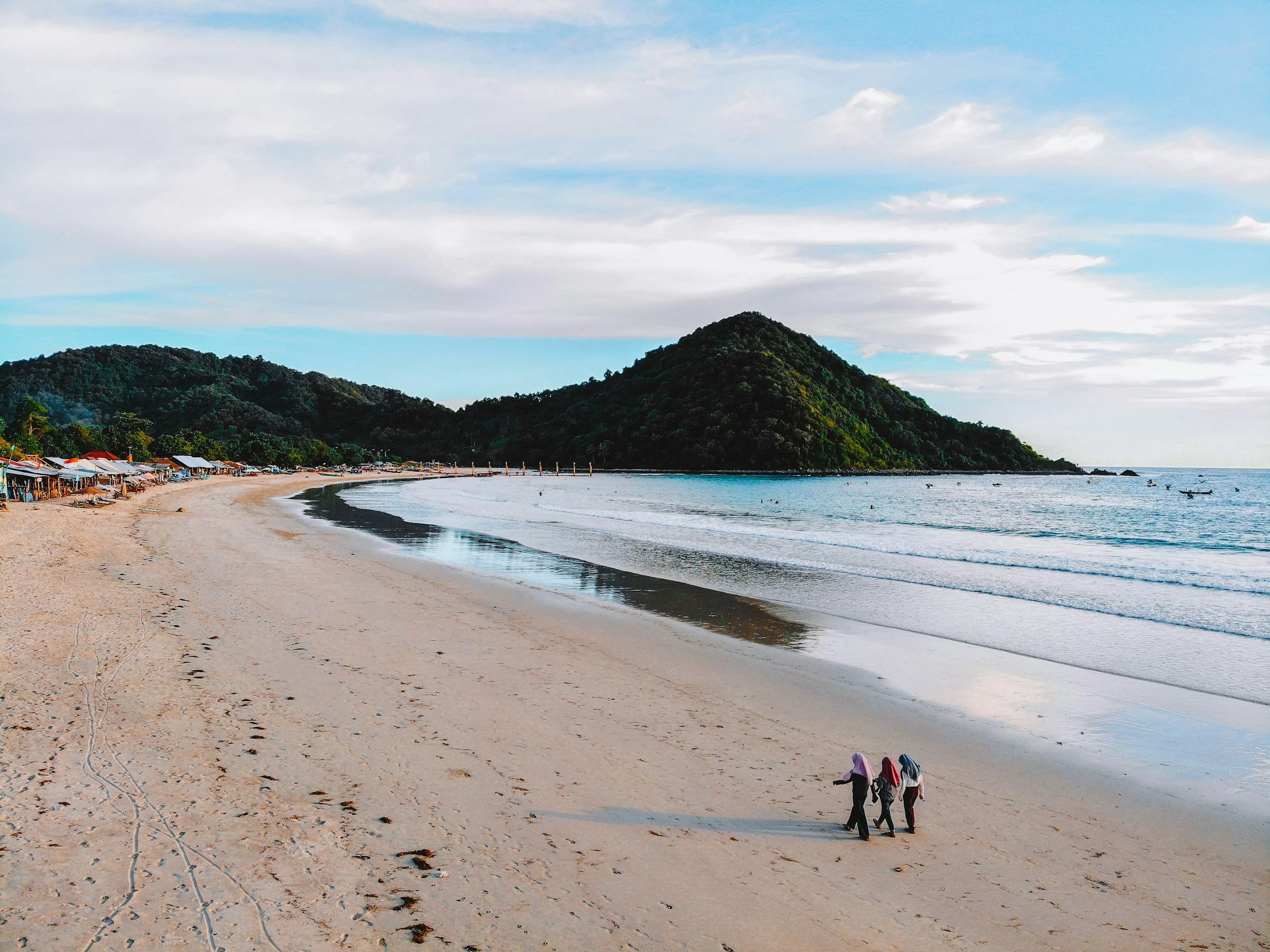 A beach with footprints and tracks in the sand, small huts along the shoreline, three people walking with head coverings, a mountain in the background, and boats on the water under a partly cloudy sky.