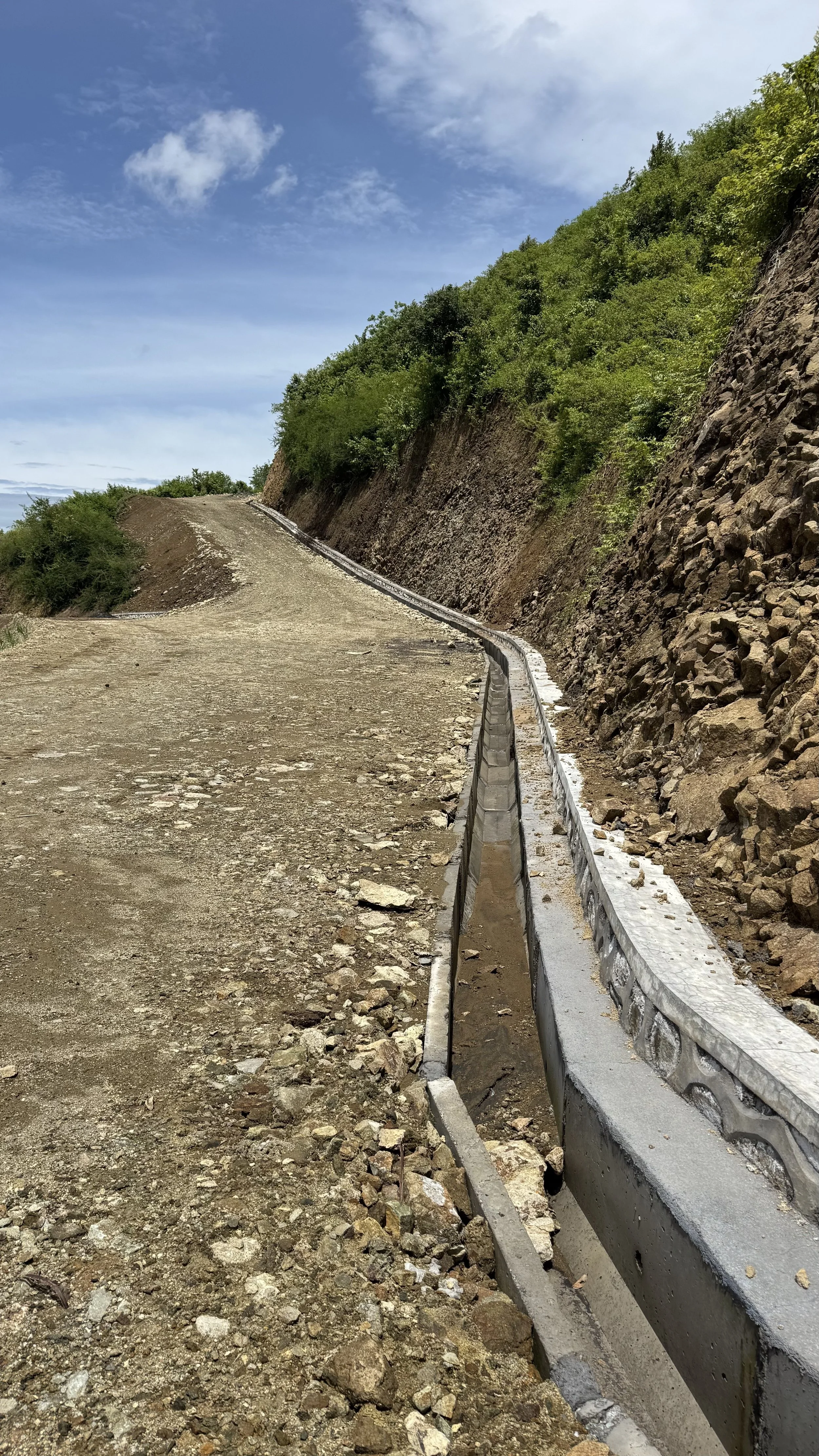 A mountain road under construction with a temporary barrier on the right side, steep rocky hillside on the right, green trees on the hillside, blue sky with scattered clouds.