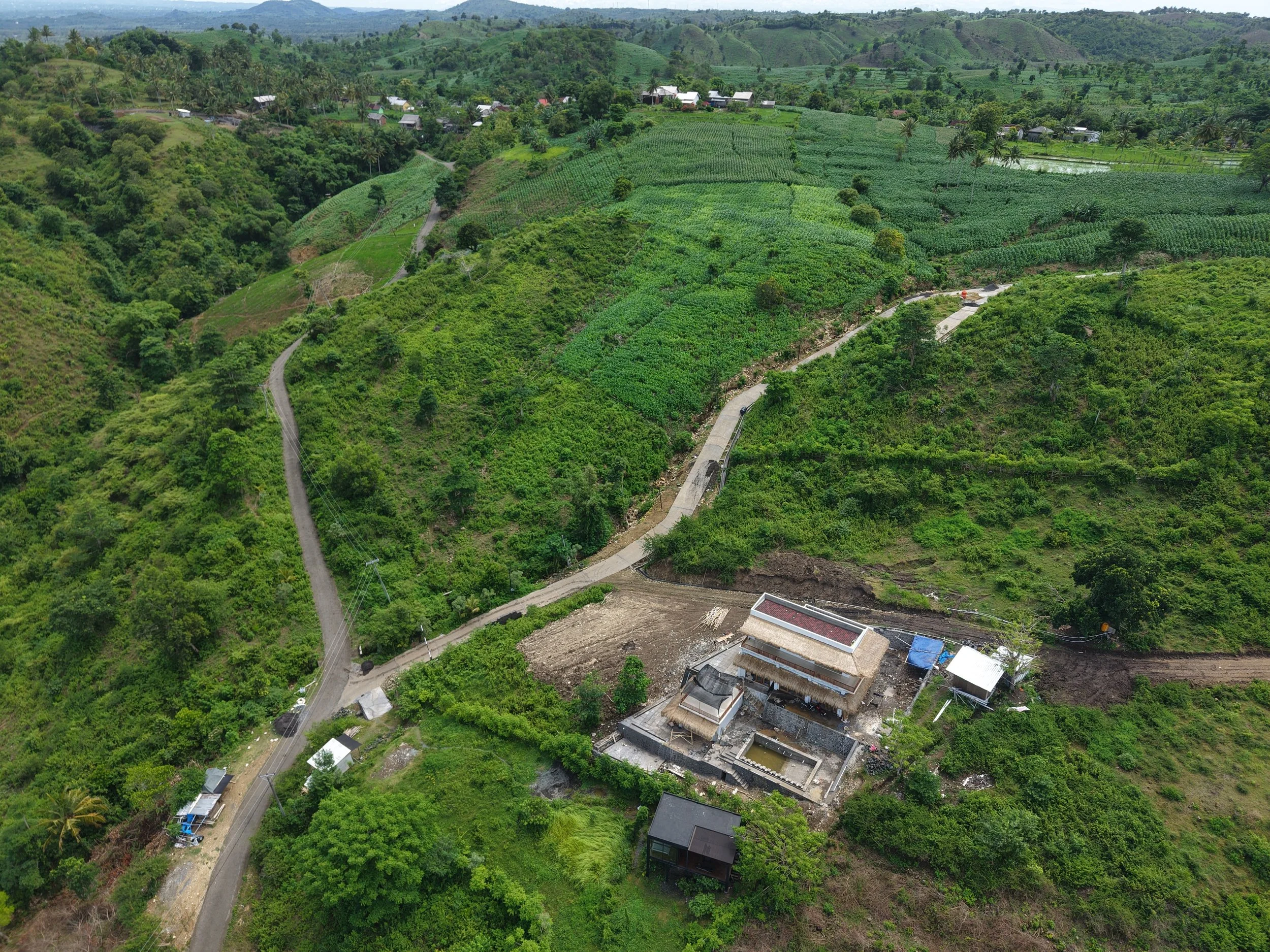 A rural hillside landscape with winding roads, green fields, trees, and a few buildings, including a large house with a red roof and a swimming pool.