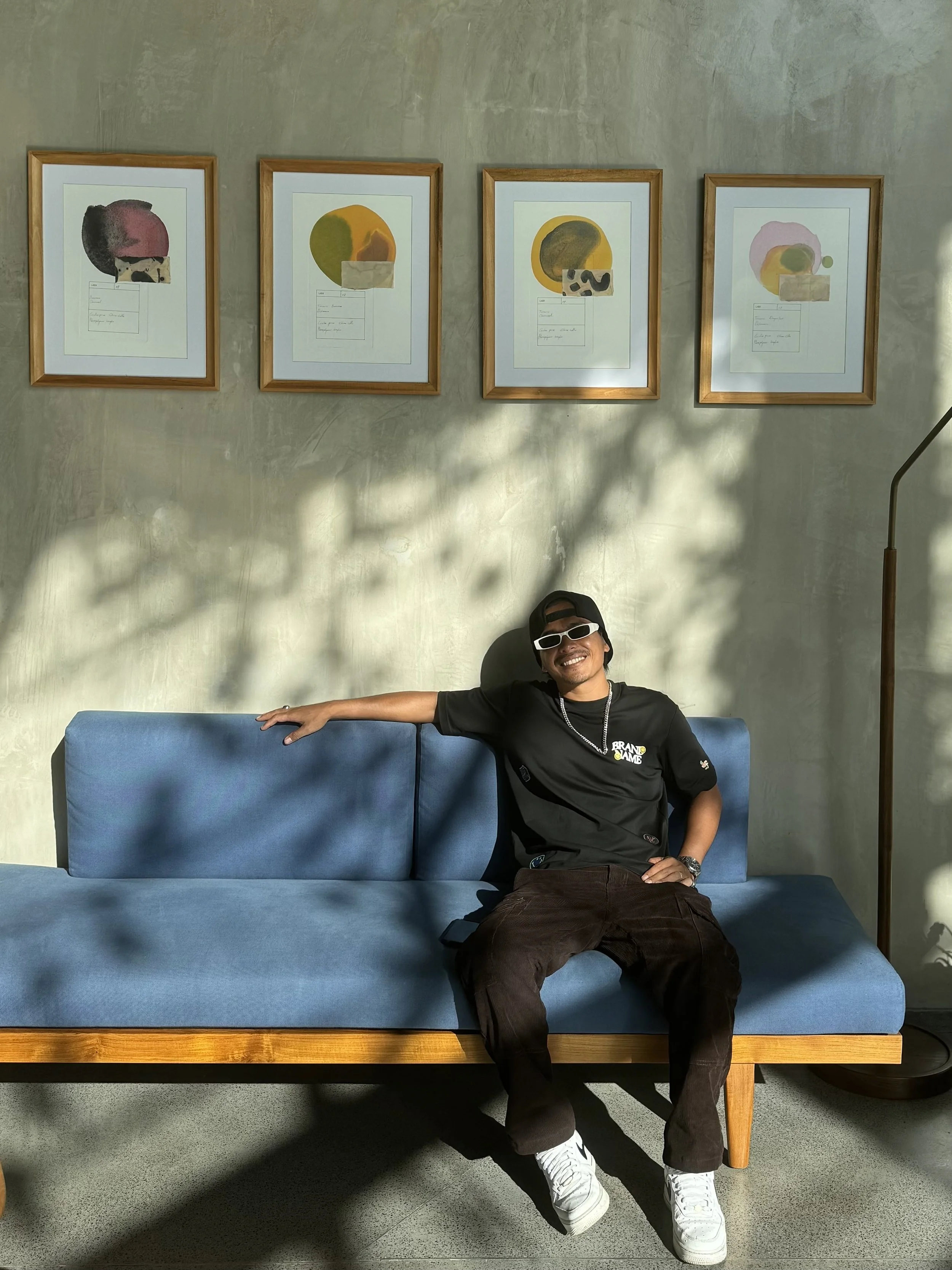 A young man wearing sunglasses, a black cap, and a black t-shirt with yellow and white text, sitting on a blue sofa with one arm resting on the back, in a room with artwork on the wall and a floor lamp nearby.