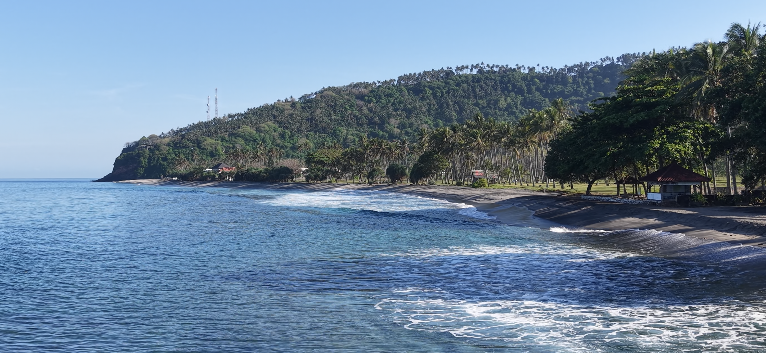 A scenic beach with clear blue water, gentle waves, and a shoreline lined with palm trees and greenery, with a hilly background.