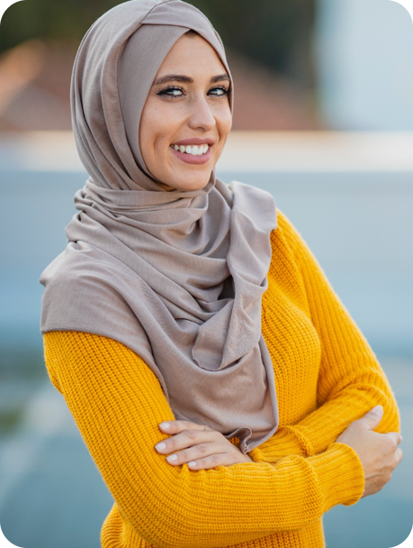 Smiling woman wearing a hijab and a yellow sweater with arms crossed outdoors.