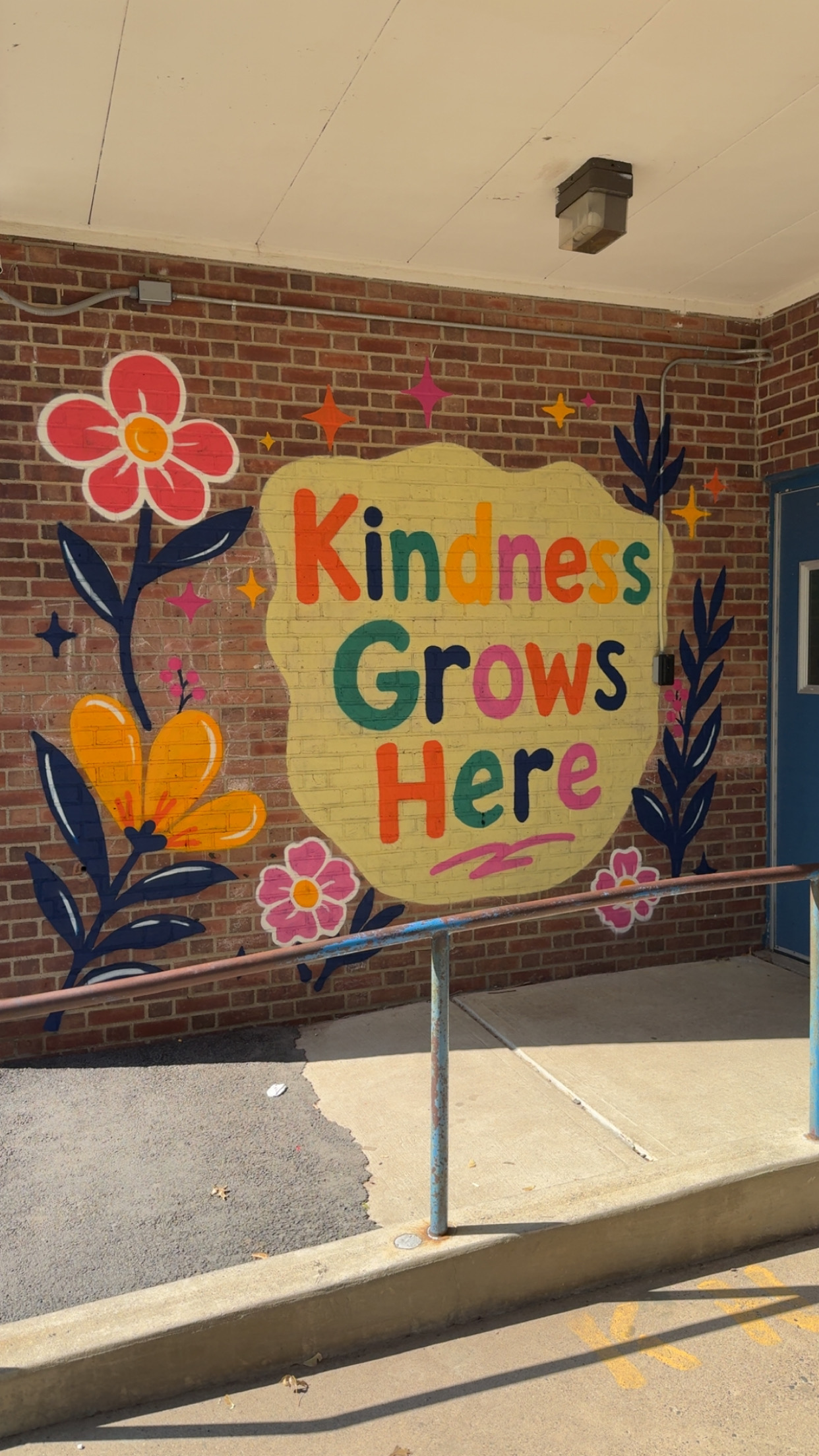 Colorful school mural painted on a brick wall with flowers, stars, and the phrase “Kindness Grows Here.”