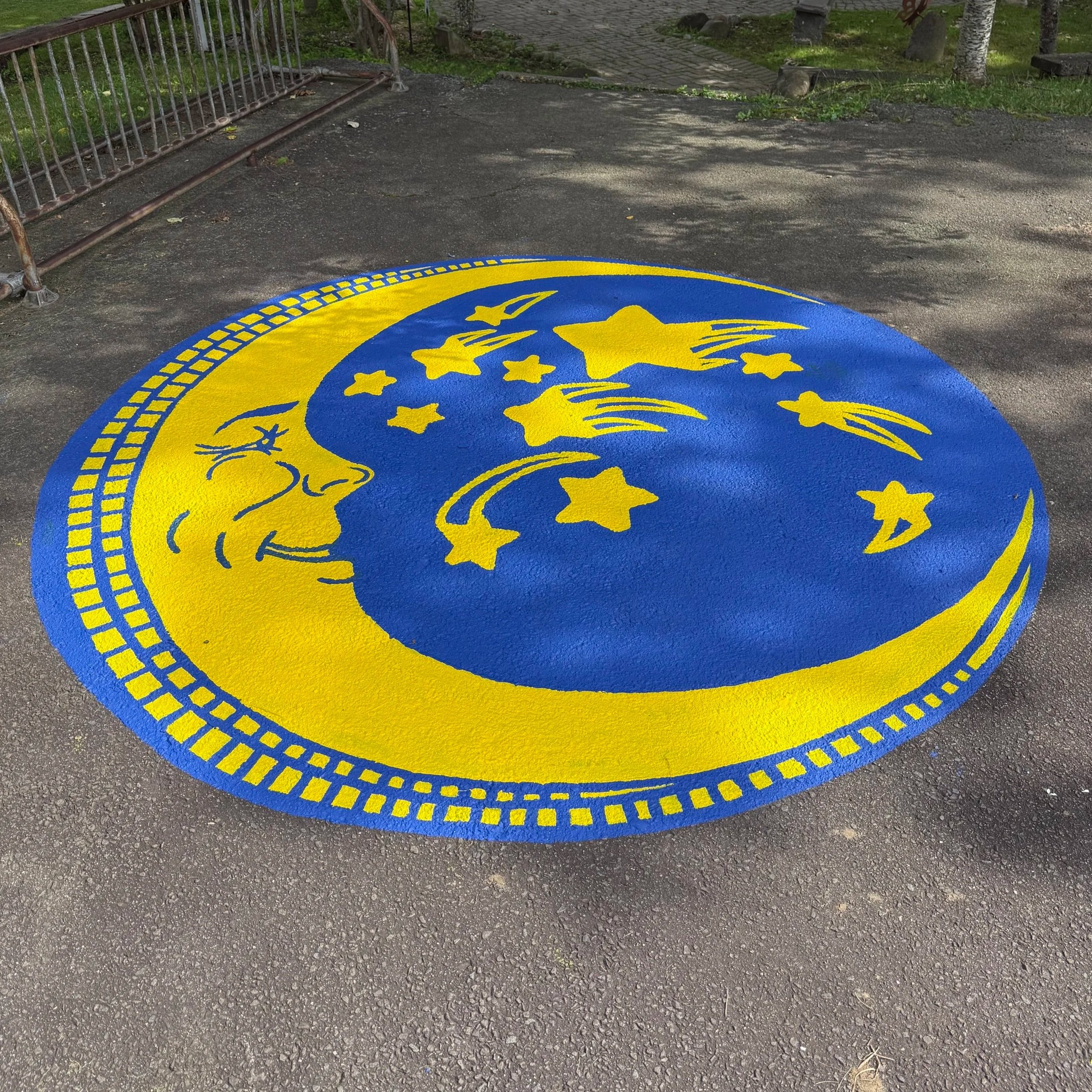 Hand-painted school pavement mural featuring a yellow moon and stars on a circular blue background.