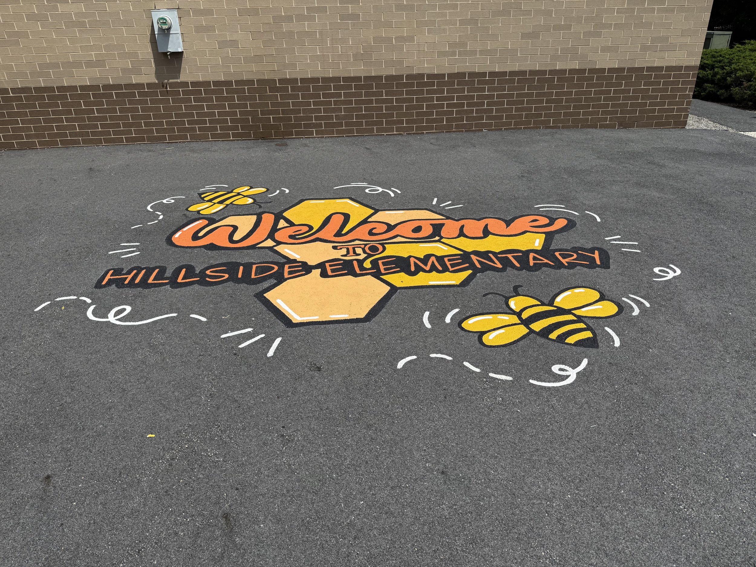 welcome sign painted on asphalt, with bees and honeycomb designs, outside Hillside Elementary School.