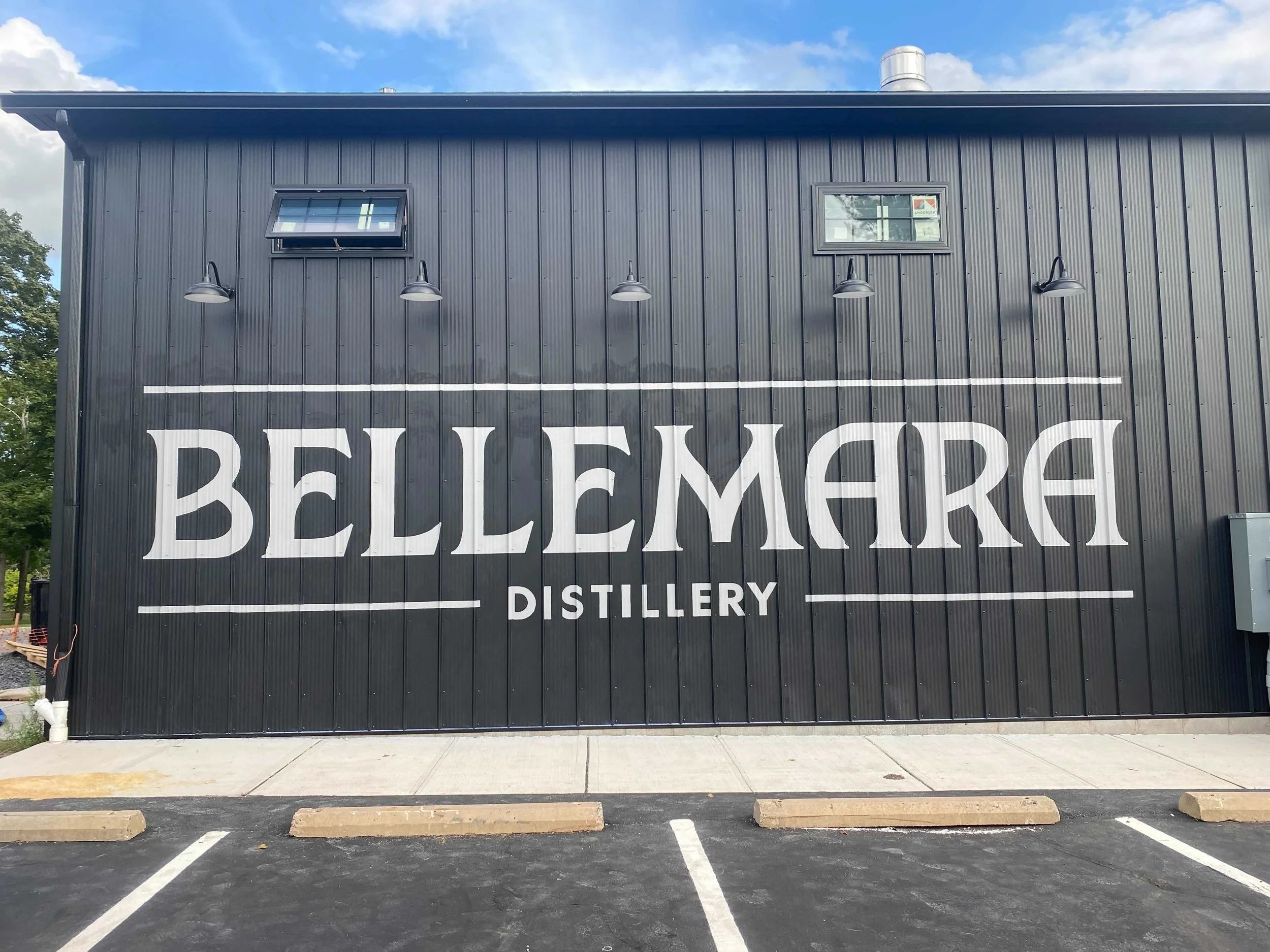 Black metal building with two small windows and four outdoor wall-mounted lights. Partial large white text on the building reading 'BELLEMARE' and 'DISTILLERY'. Empty parking lot in front.