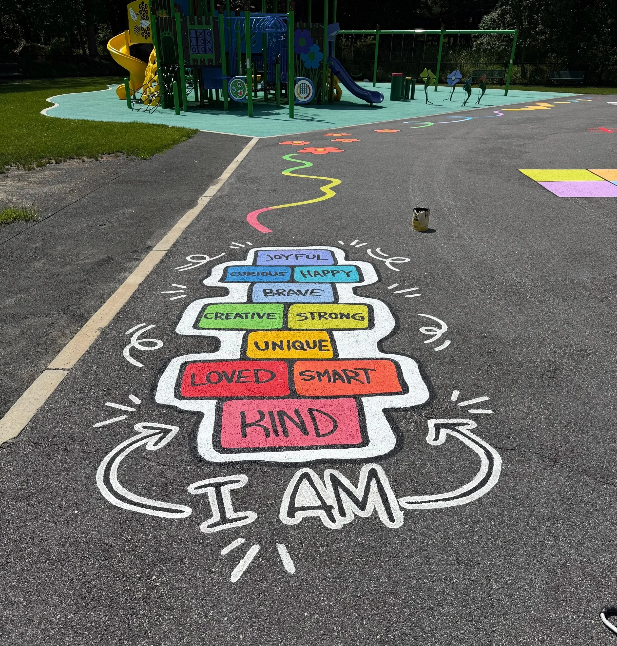 Colorful hopscotch pattern painted on pavement with words like 'Joyful,' 'Curious,' and 'Happy,' and a playground with slides, climbing structures, and painted flowers in the background.