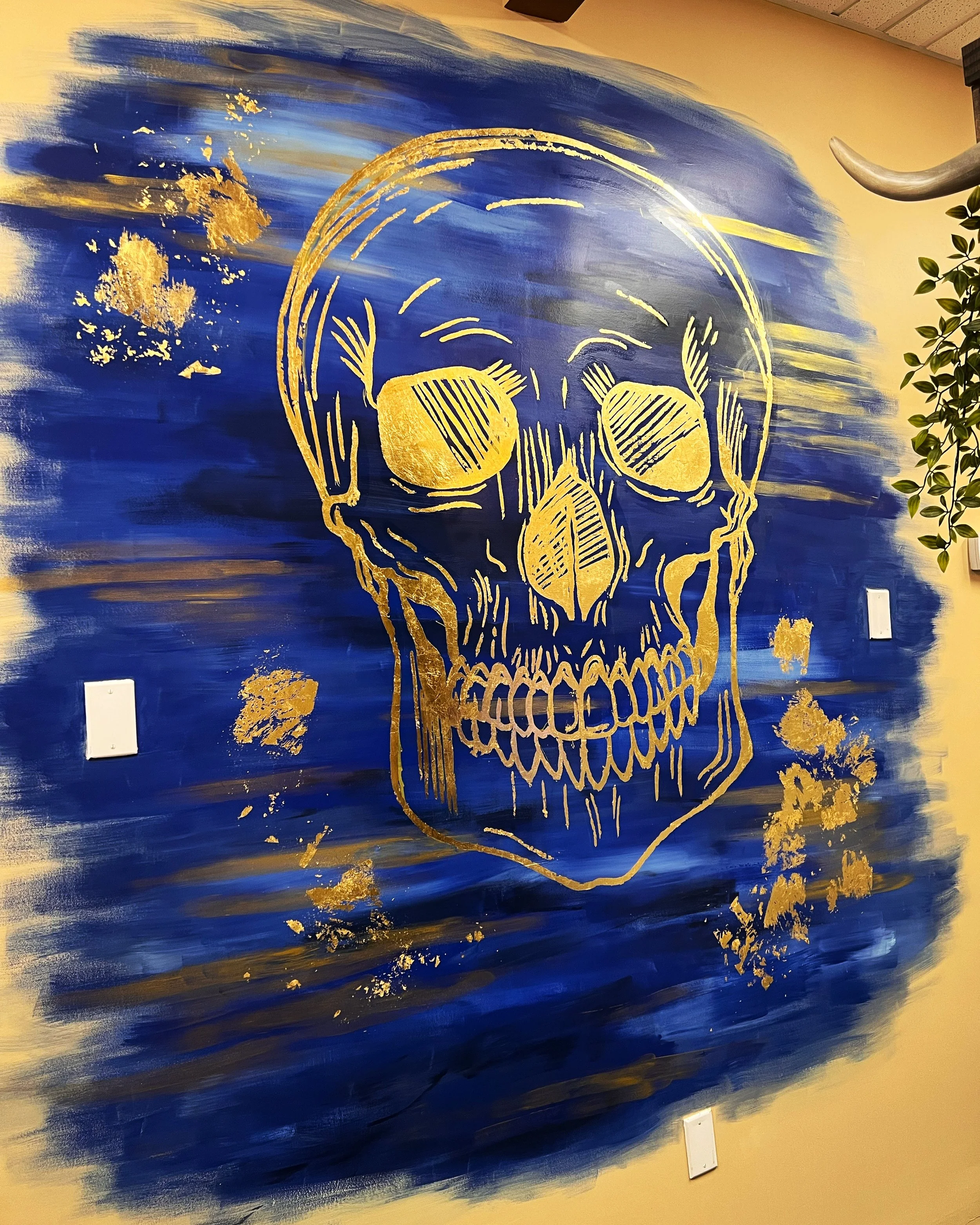 A mural of a human skull outlined in gold on a blue and gold abstract background, located on a beige wall inside a building with ceiling tiles and a ceiling vent.