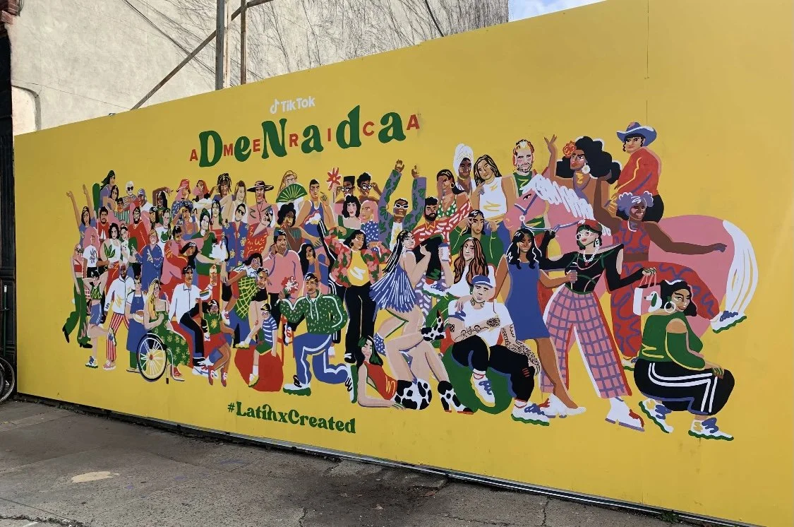 Colorful mural of diverse people celebrating Latin culture, including dancers, musicians, and community members, on a bright yellow wall with the hashtag #LatinhxCreated.