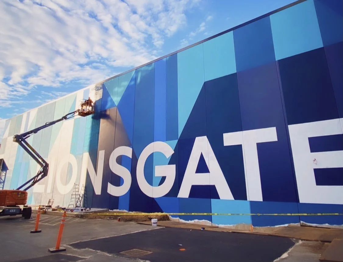 Construction worker on a lift painting a large mural on a building wall, with the letters 'LIONSGATE' partly visible.
