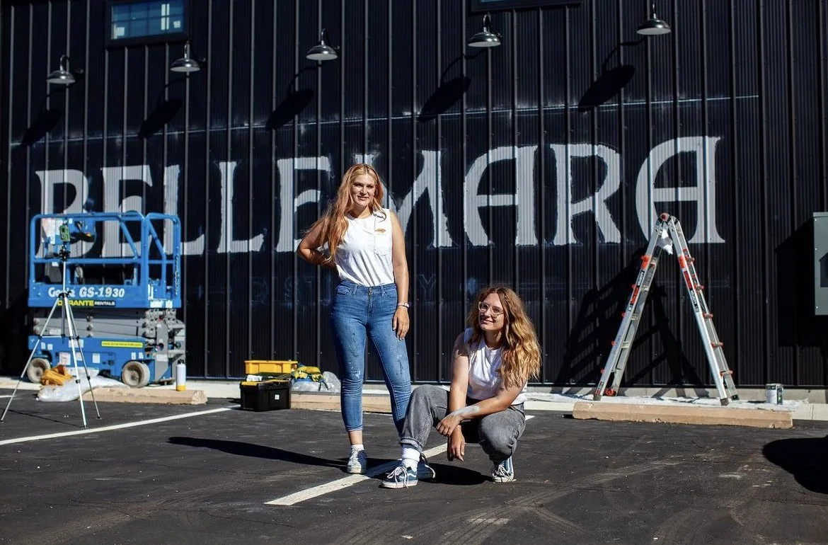 Two women with long, wavy hair, one standing and one squatting, in front of a black building with large white signs, yard equipment, a ladder, and construction tools around.