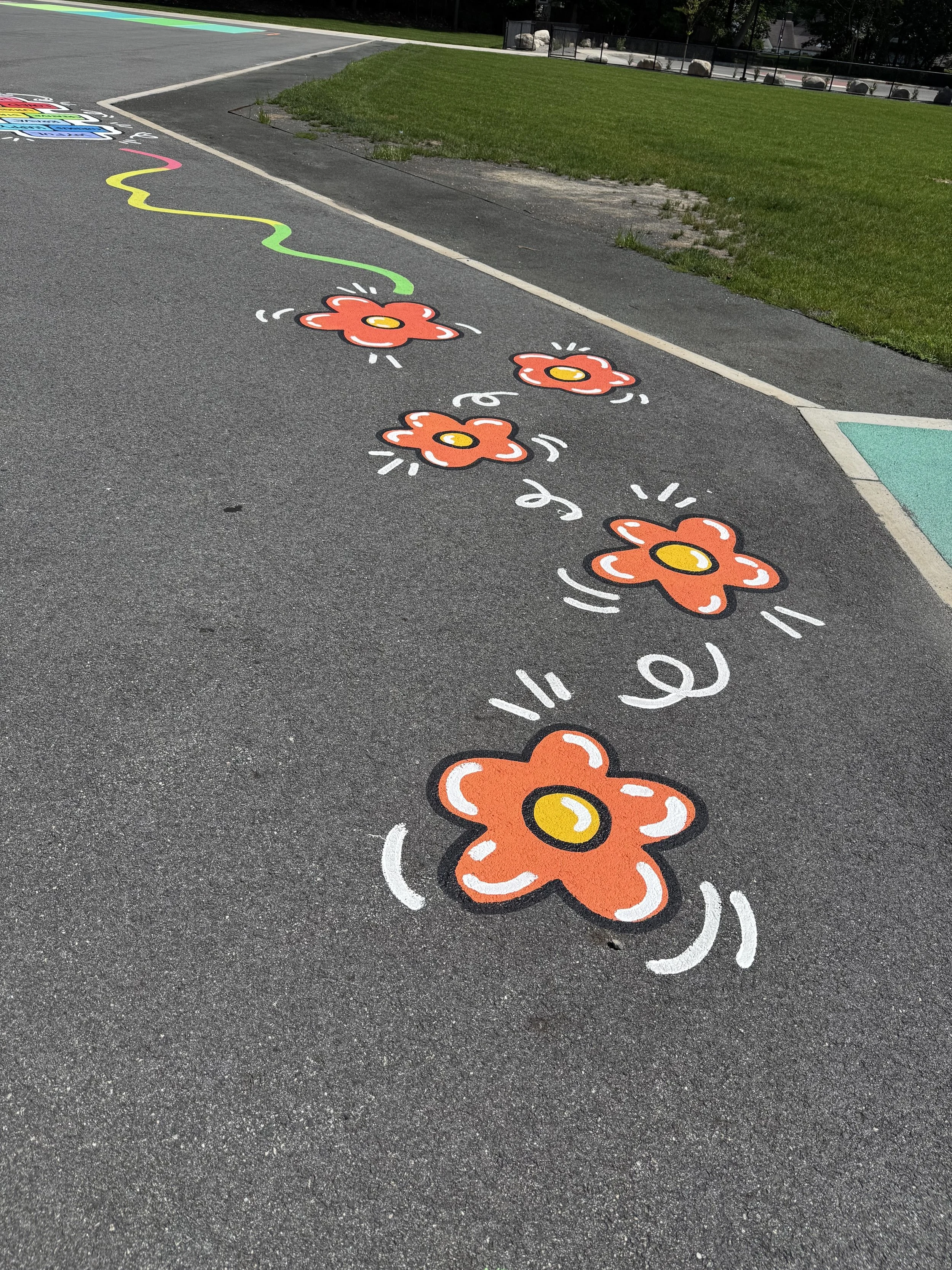 Outdoor school mural with colorful painted flowers and a rainbow pathway on black pavement.
