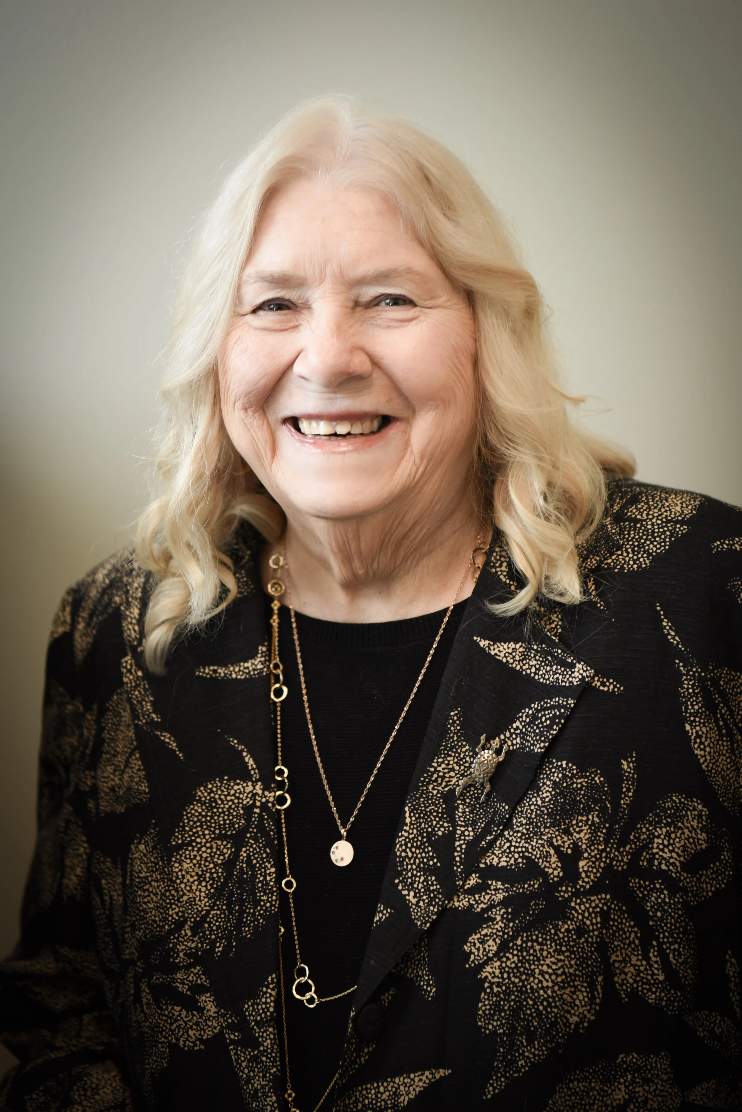 Smiling elderly woman with blonde hair wearing a black and red patterned top and a silver necklace.