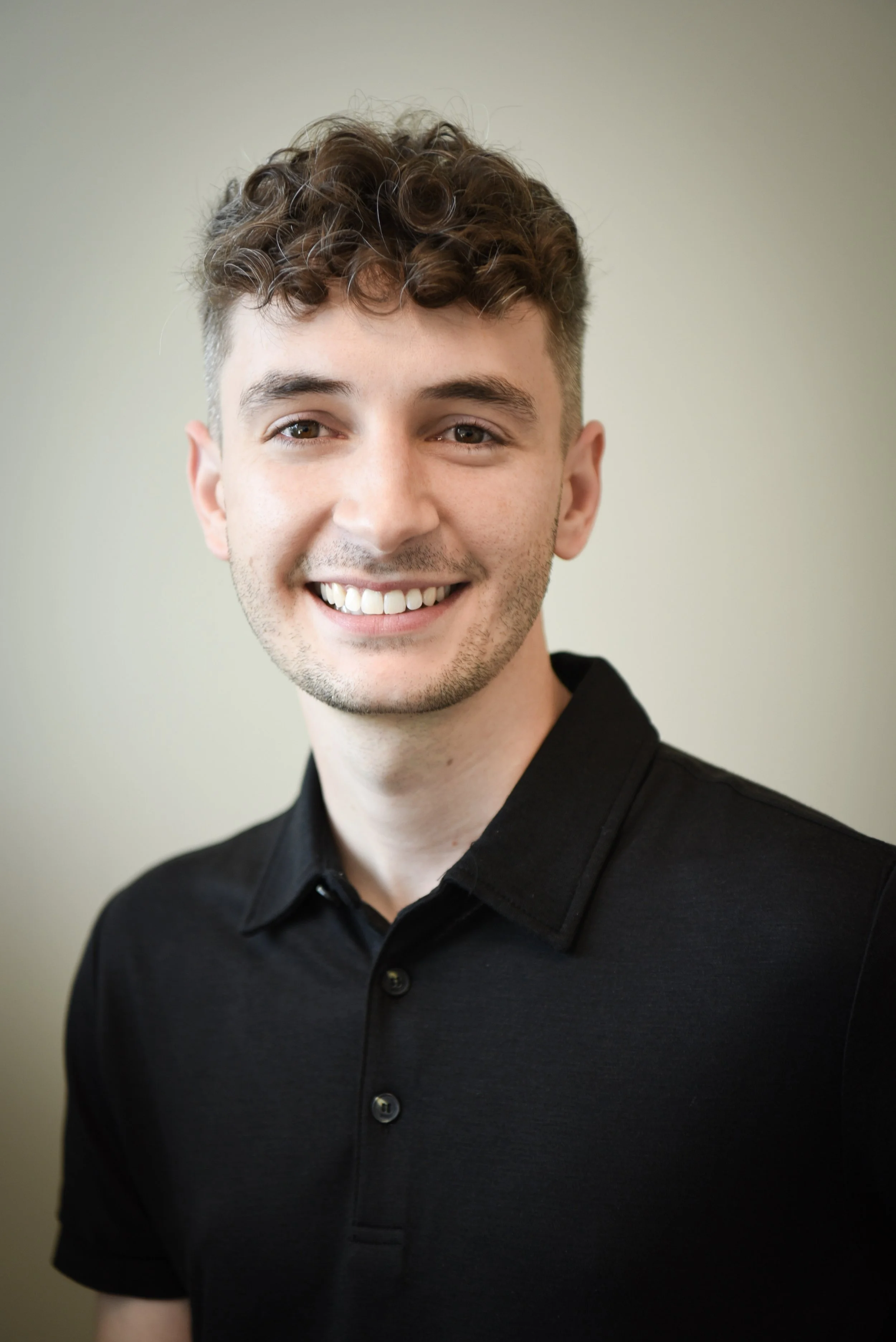 A young man with short, curly brown hair and a light skin tone smiling, wearing a black collared shirt, against a plain neutral background.