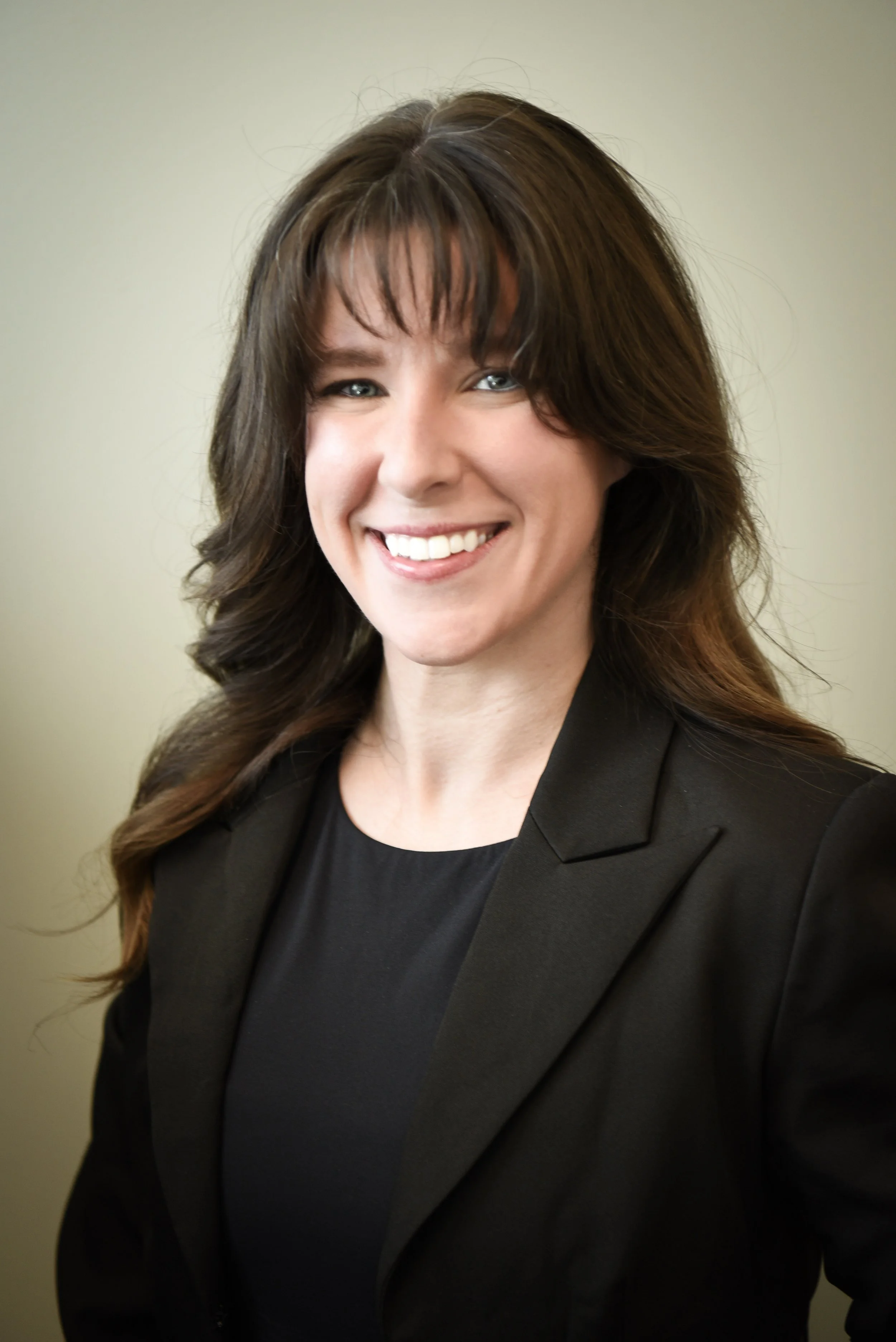 Smiling woman with glasses and a necklace, sitting in a room with a bookshelf and window in the background.