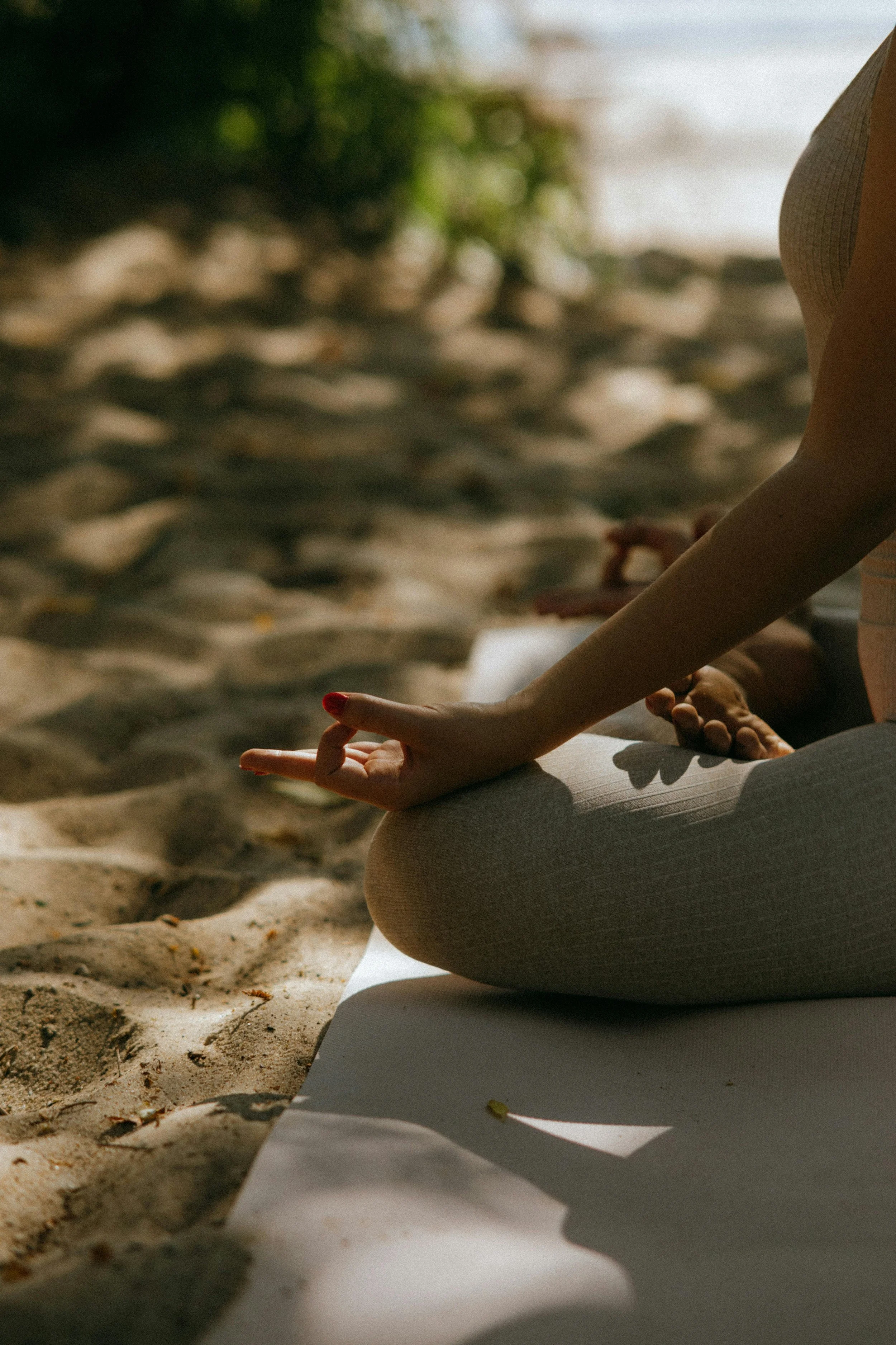A person practicing yoga or meditation on a beach, sitting cross-legged on a mat with hand gestures, sand and trees in the background.